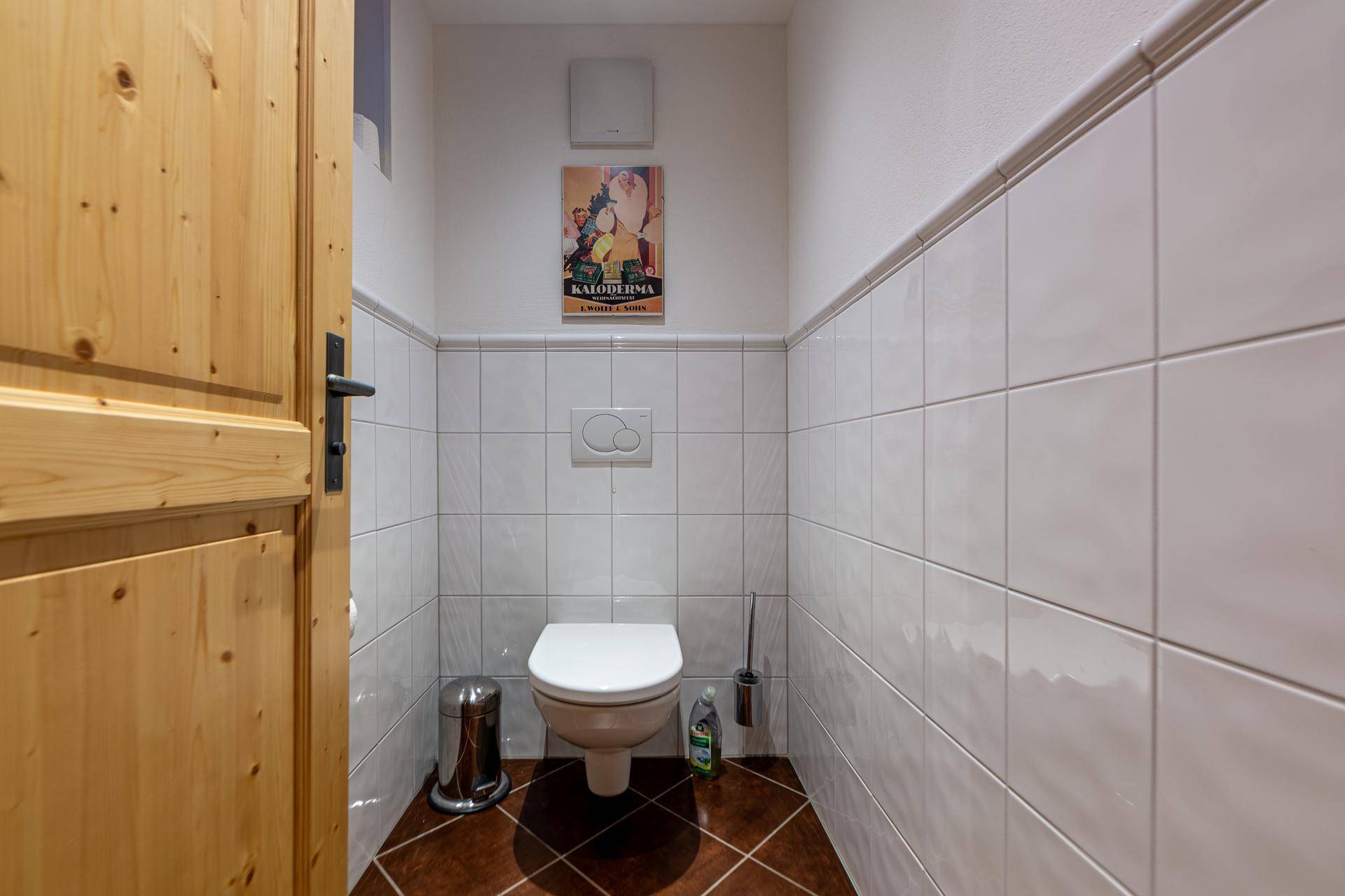 Separate Toilette mit Wand-WC und weißen Fliesen, sowie braunem Fliesenboden.