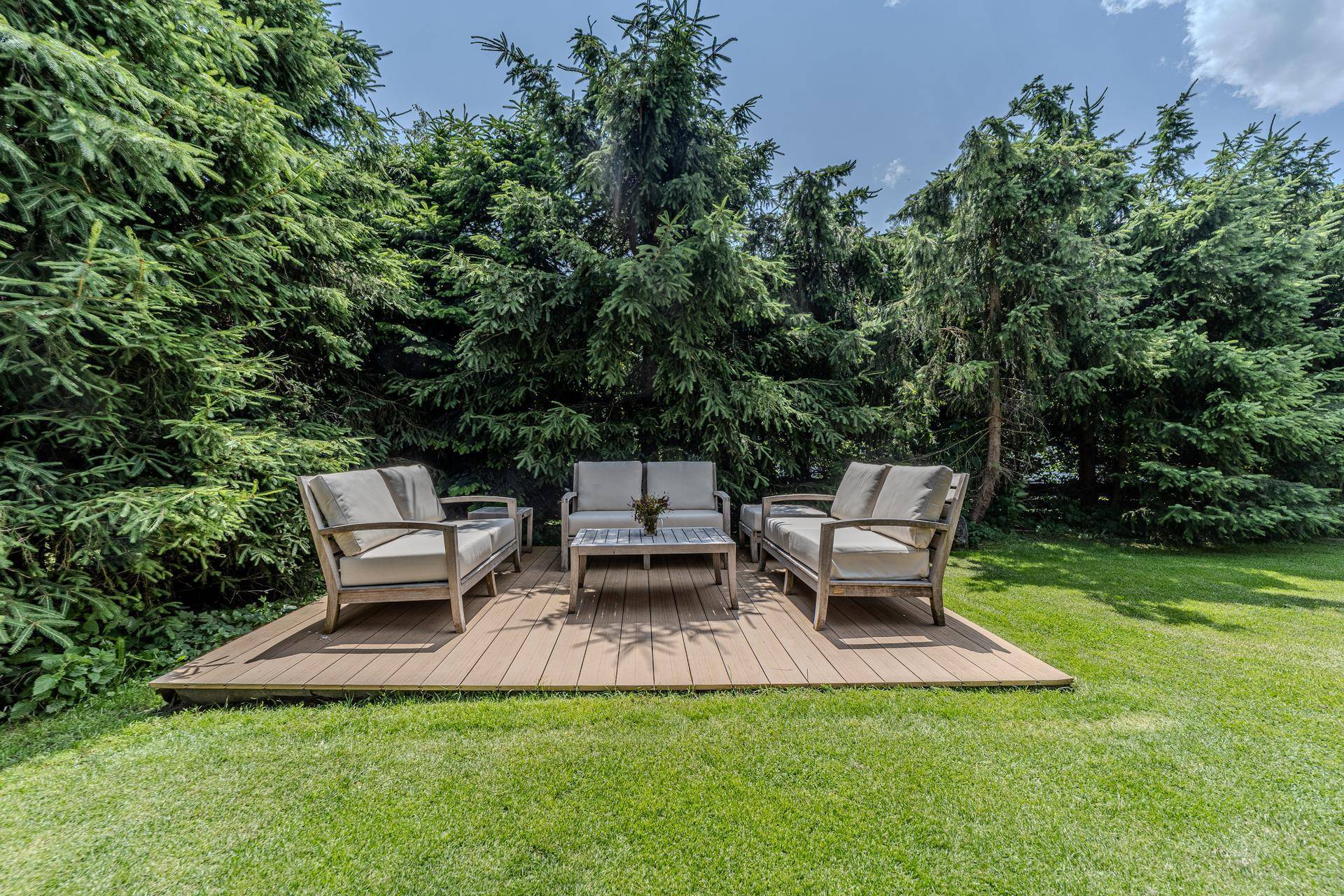 Entspannungsbereich im Garten mit bequemen Lounge-Möbeln auf einem Holzdeck, umgeben von hohen Bäumen.