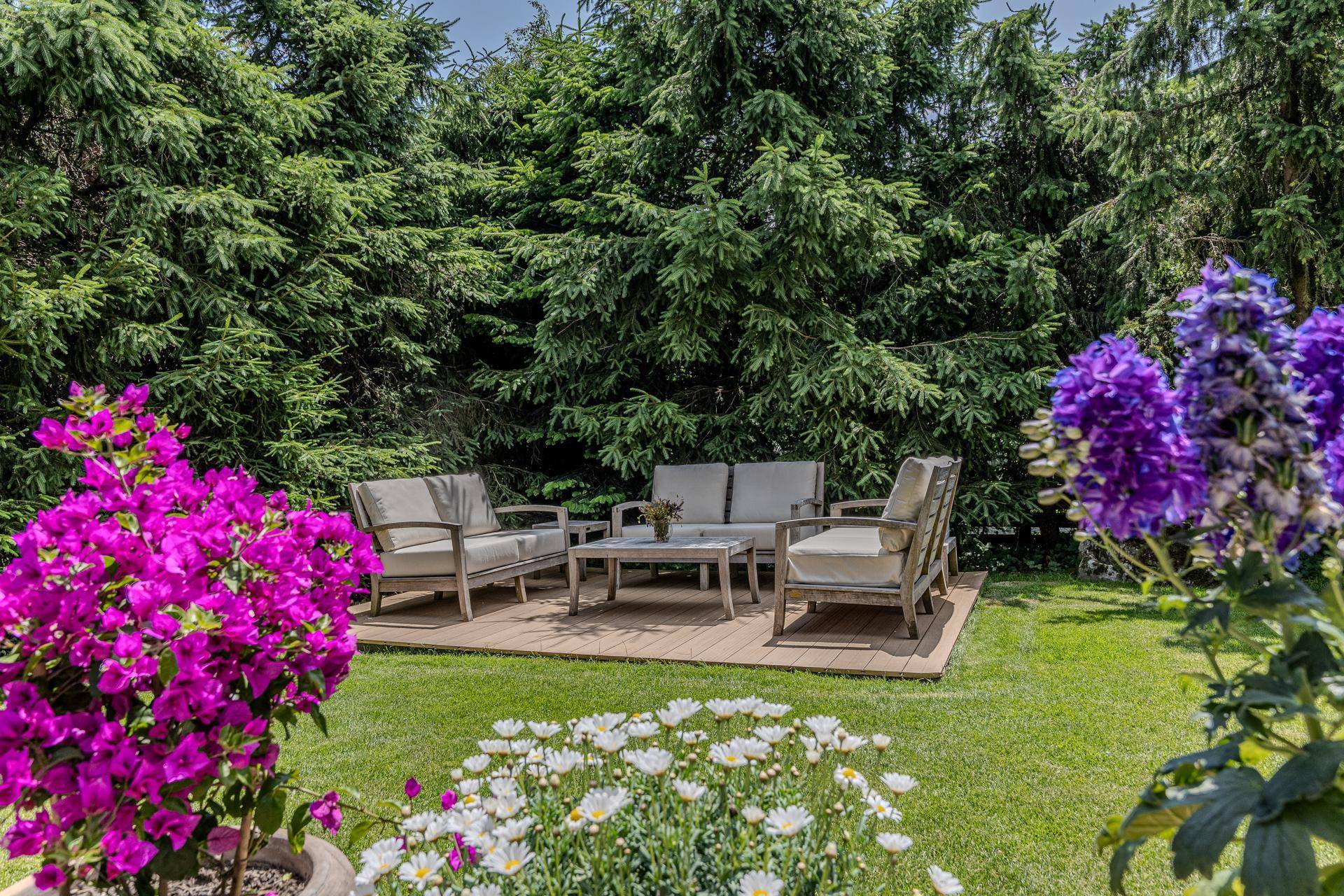 Gemütlicher Gartenbereich mit Lounge-Möbeln auf einem Holzdeck, umrahmt von hohen Nadelbäumen und bunten Blumen.