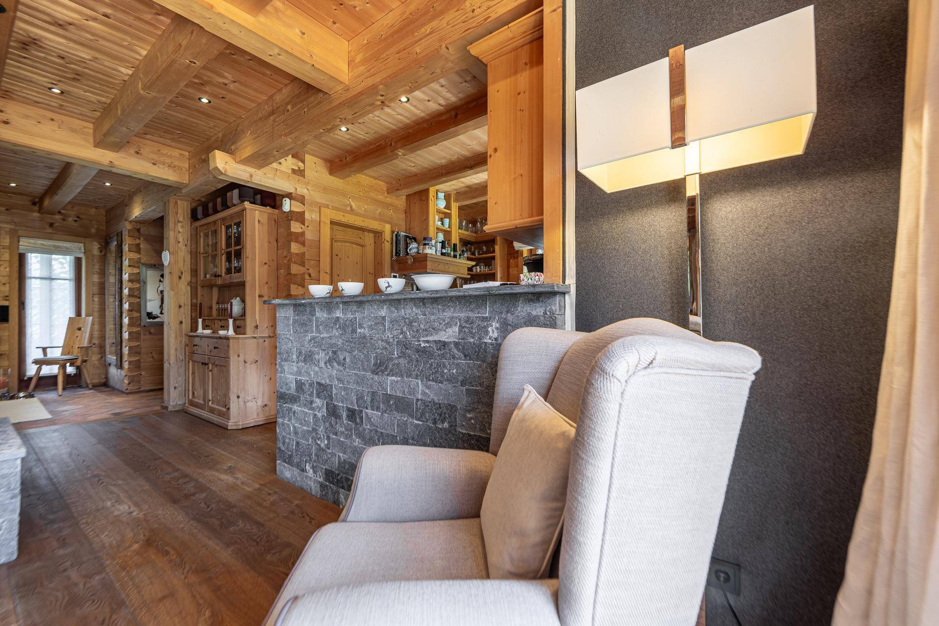 Gemütlicher Wohnbereich mit Sessel und Steinwand, offen zur Küche und mit Holzbalkendecke.