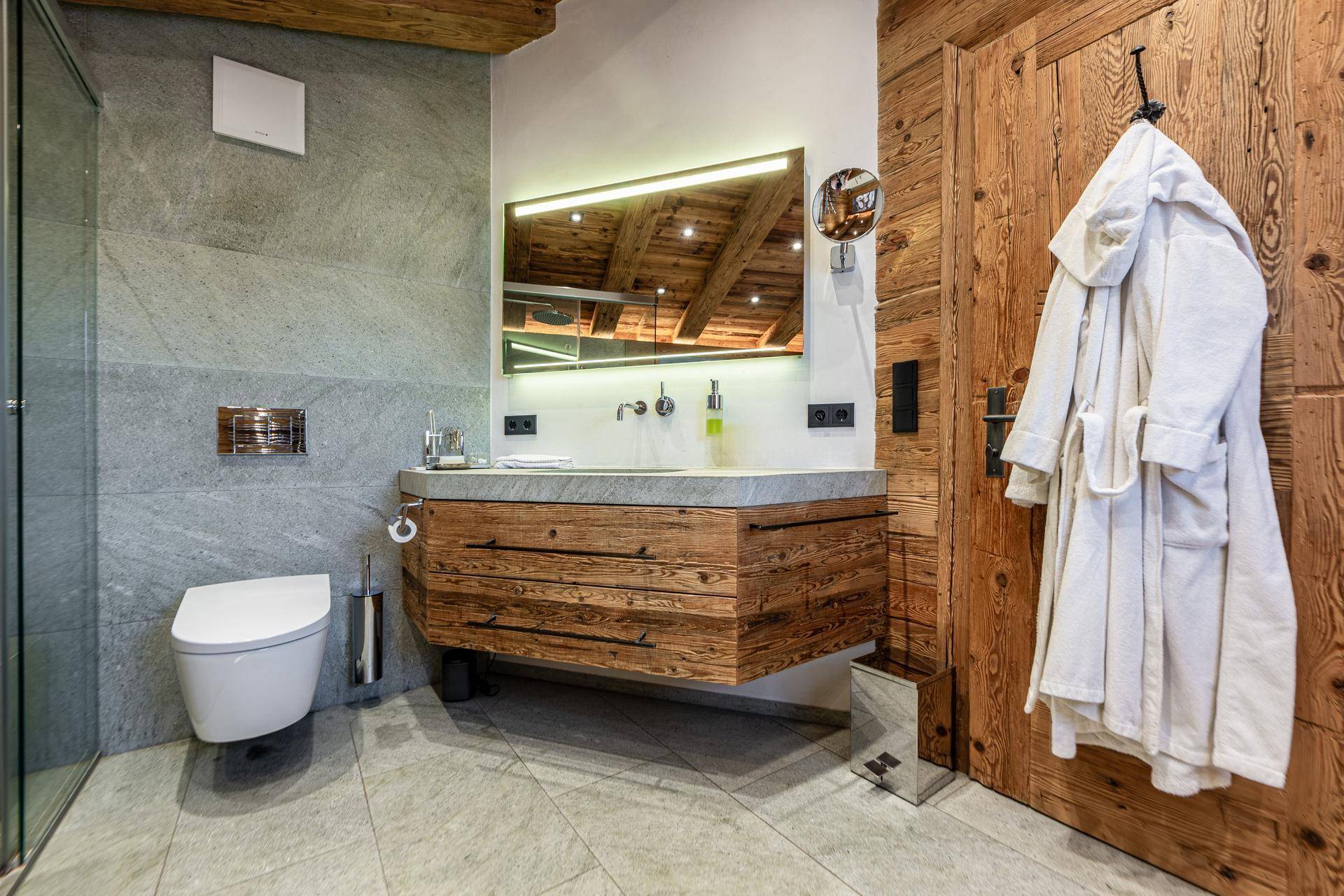 Elegantes Badezimmer mit Holzwaschtisch, Wand-WC und großem Spiegel mit integrierter Beleuchtung.