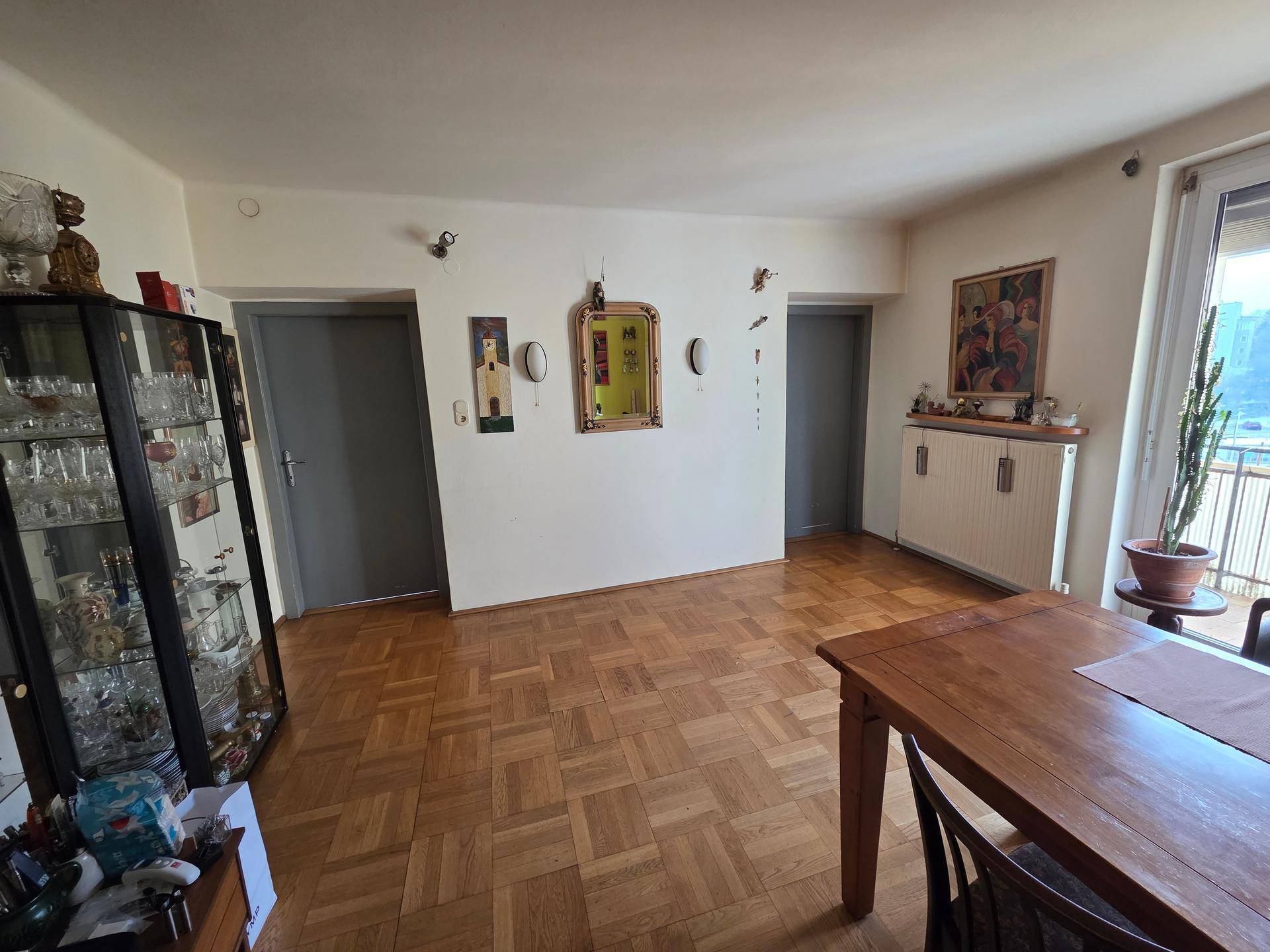 Wohn- und Essbereich mit Parkettboden, Vitrine und Zugang zum Balkon.