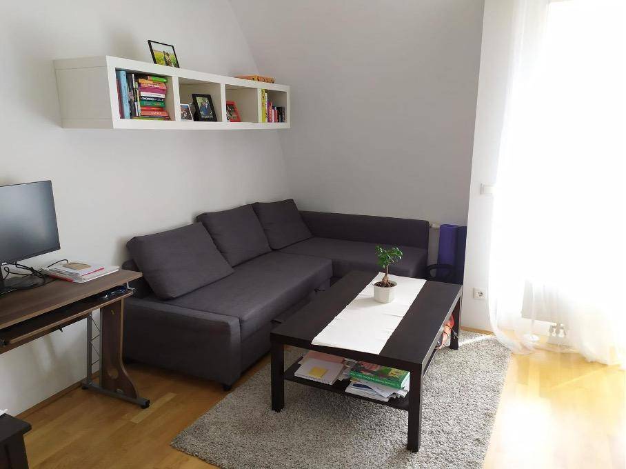 Gemütliches Wohnzimmer mit Ecksofa, Schreibtisch und Dachschräge, hell durch großes Fenster.