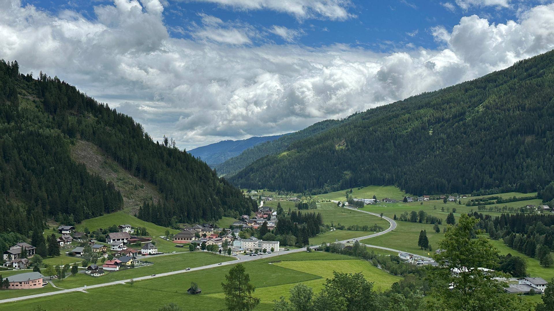 Panoramablick auf ein grünes Tal mit einem malerischen Dorf und bewaldeten Bergen unter einem bewölkten Himmel.