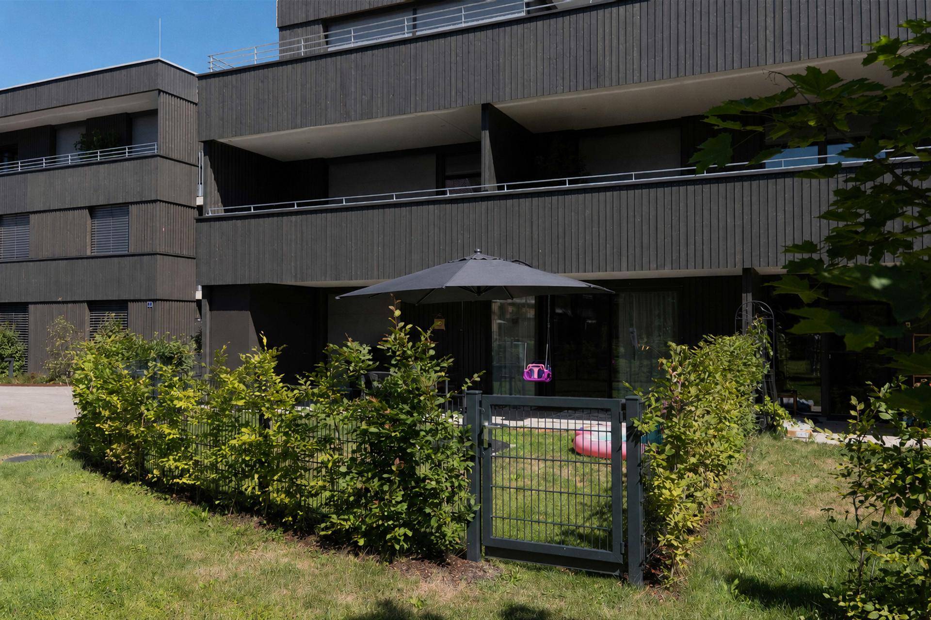 Gepflegter Gartenbereich mit Sonnenschirm und Zugang zu einem modernen Mehrfamilienhaus.