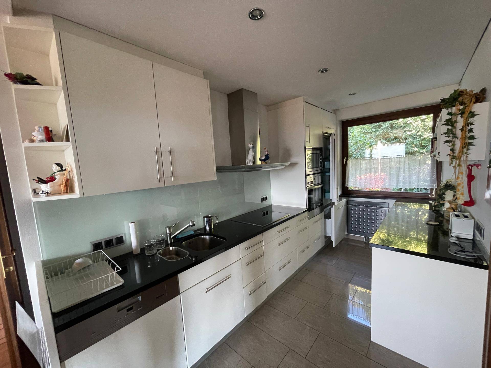 Moderne Küche mit weißen Schränken, schwarzen Arbeitsplatten, Fliesenboden und Fenster.