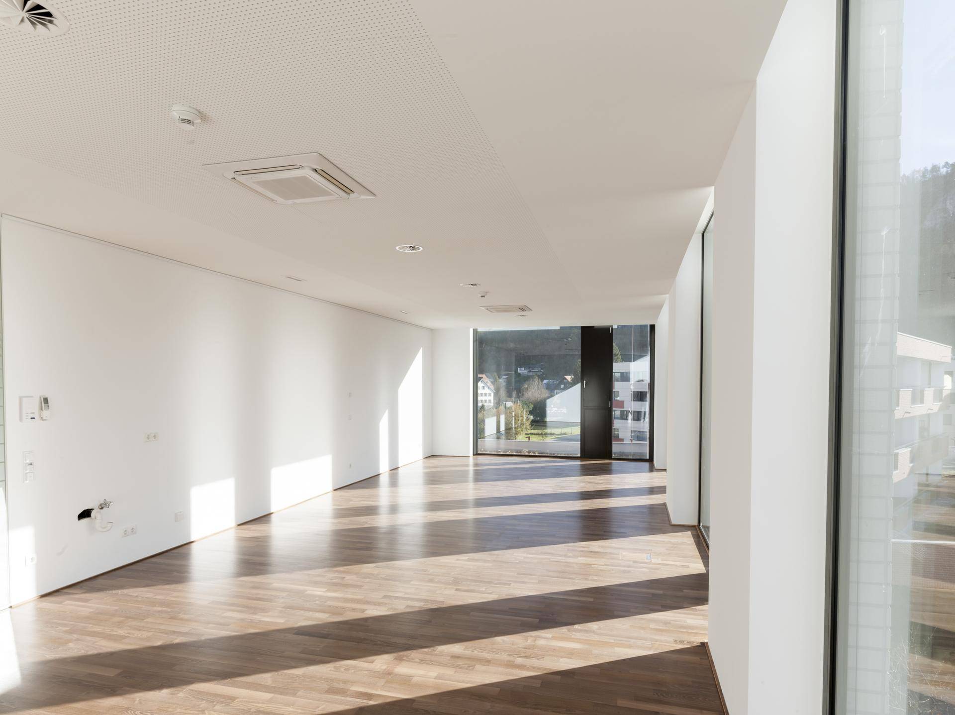 Langer, heller Büroraum mit Holzboden und raumhohen Fenstern, die den Raum mit natürlichem Licht durchfluten.