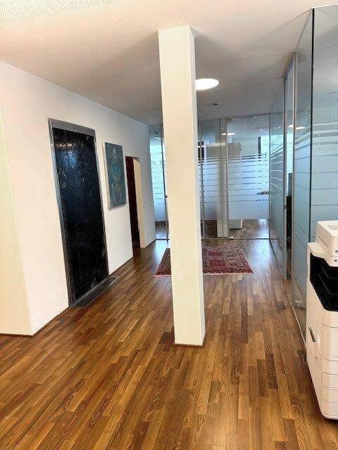 Heller Flur mit Holzboden und Glaswänden, der zu verschiedenen Büros führt und eine offene Raumgestaltung bietet.