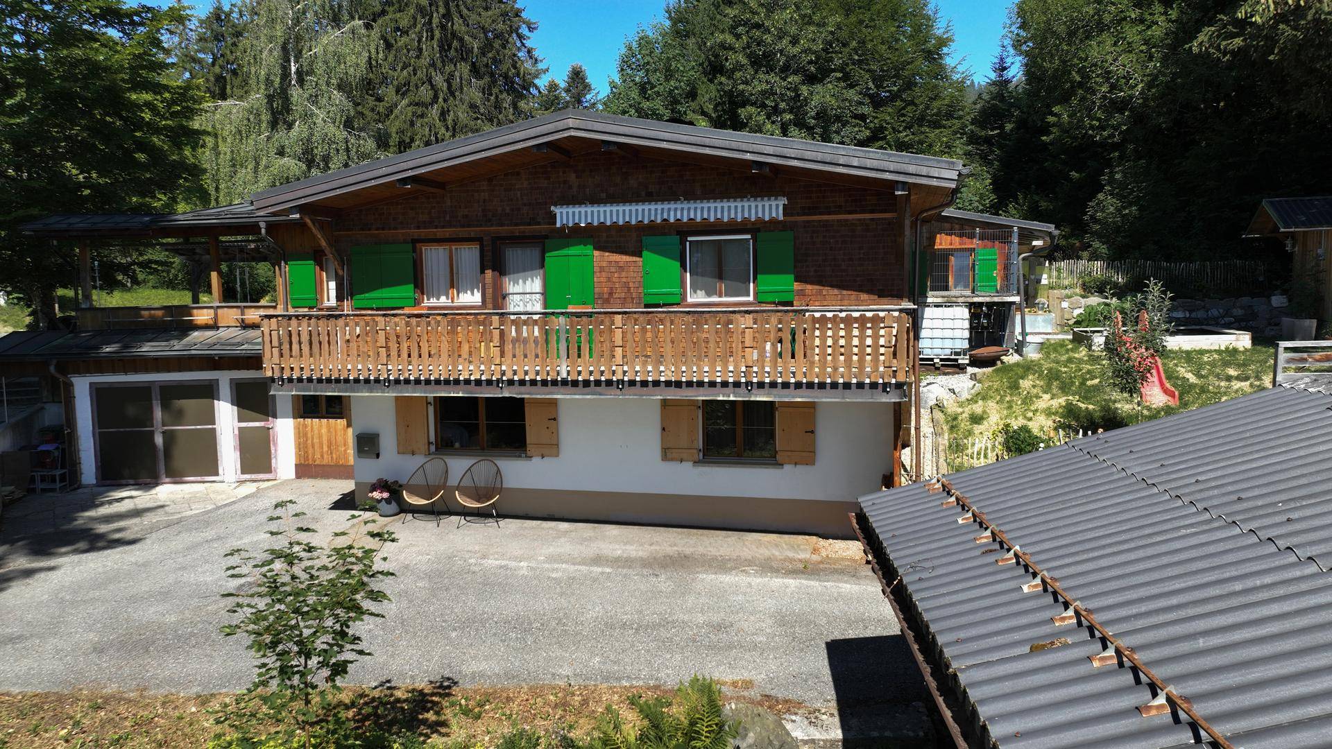 Traditionelles Holzhaus mit grünen Fensterläden und Balkon, umgeben von Bäumen und Natur.