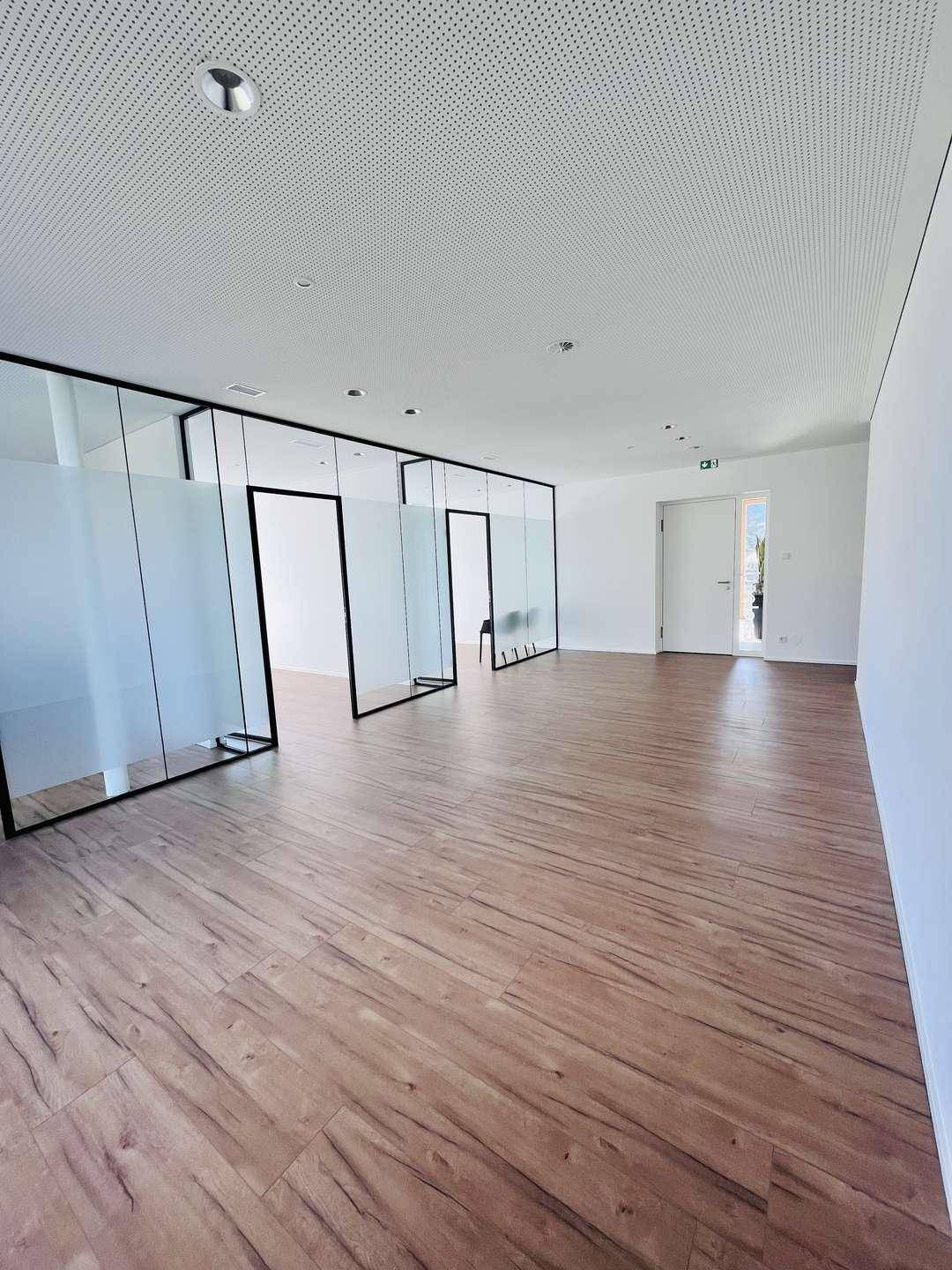 Geräumiger, heller Büroraum mit modernen Glasabtrennungen und Holzoptik-Boden.