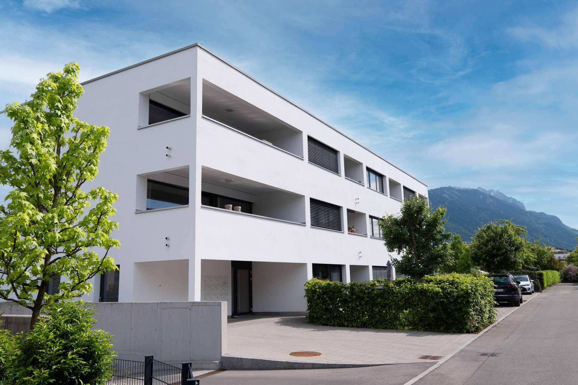 Zeitgenössisches Wohngebäude mit weißer Fassade, Balkonen und Bergblick im Hintergrund.