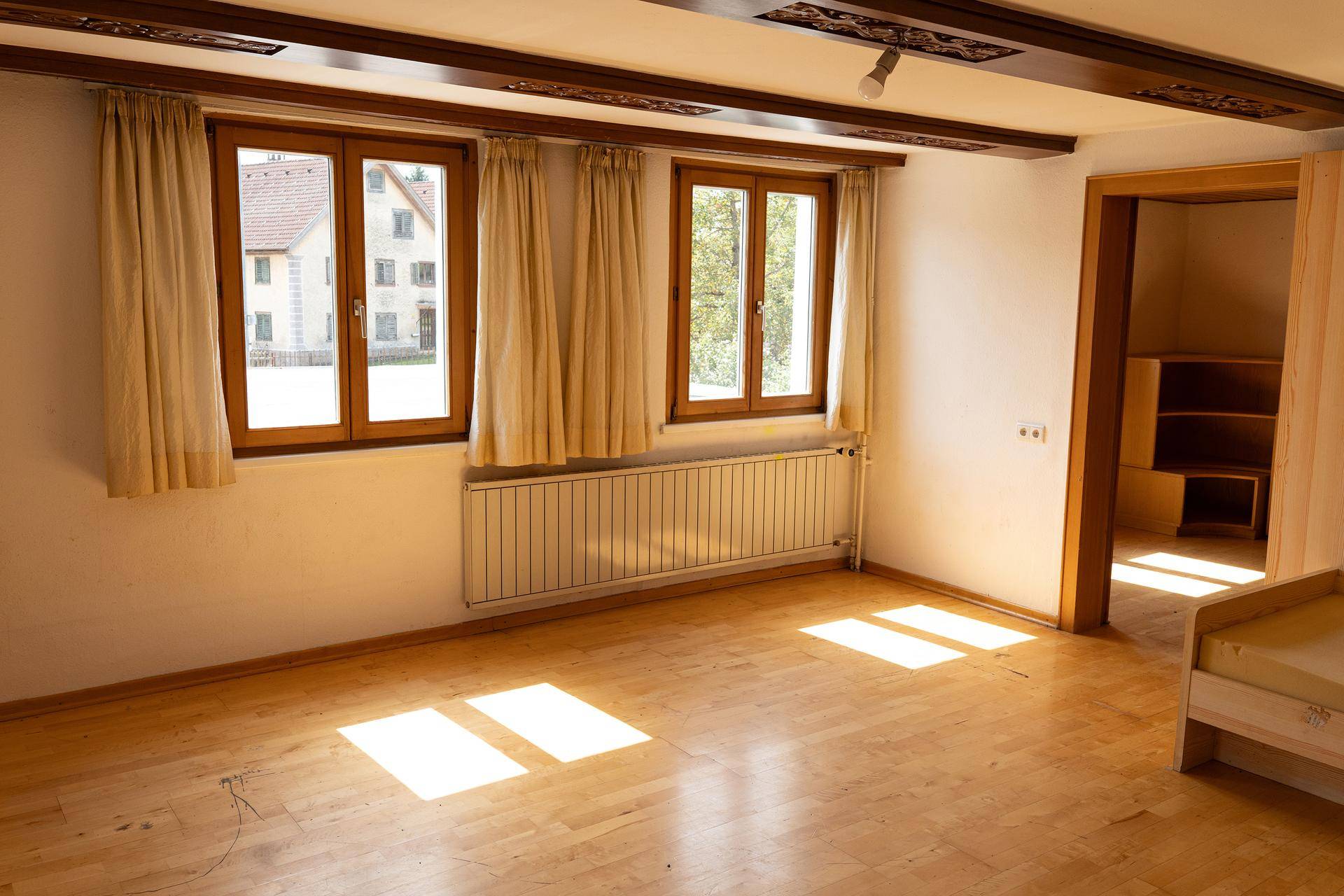 Heller Raum mit Holzboden, sichtbaren Deckenbalken und großen Fenstern, die viel Licht hereinlassen.