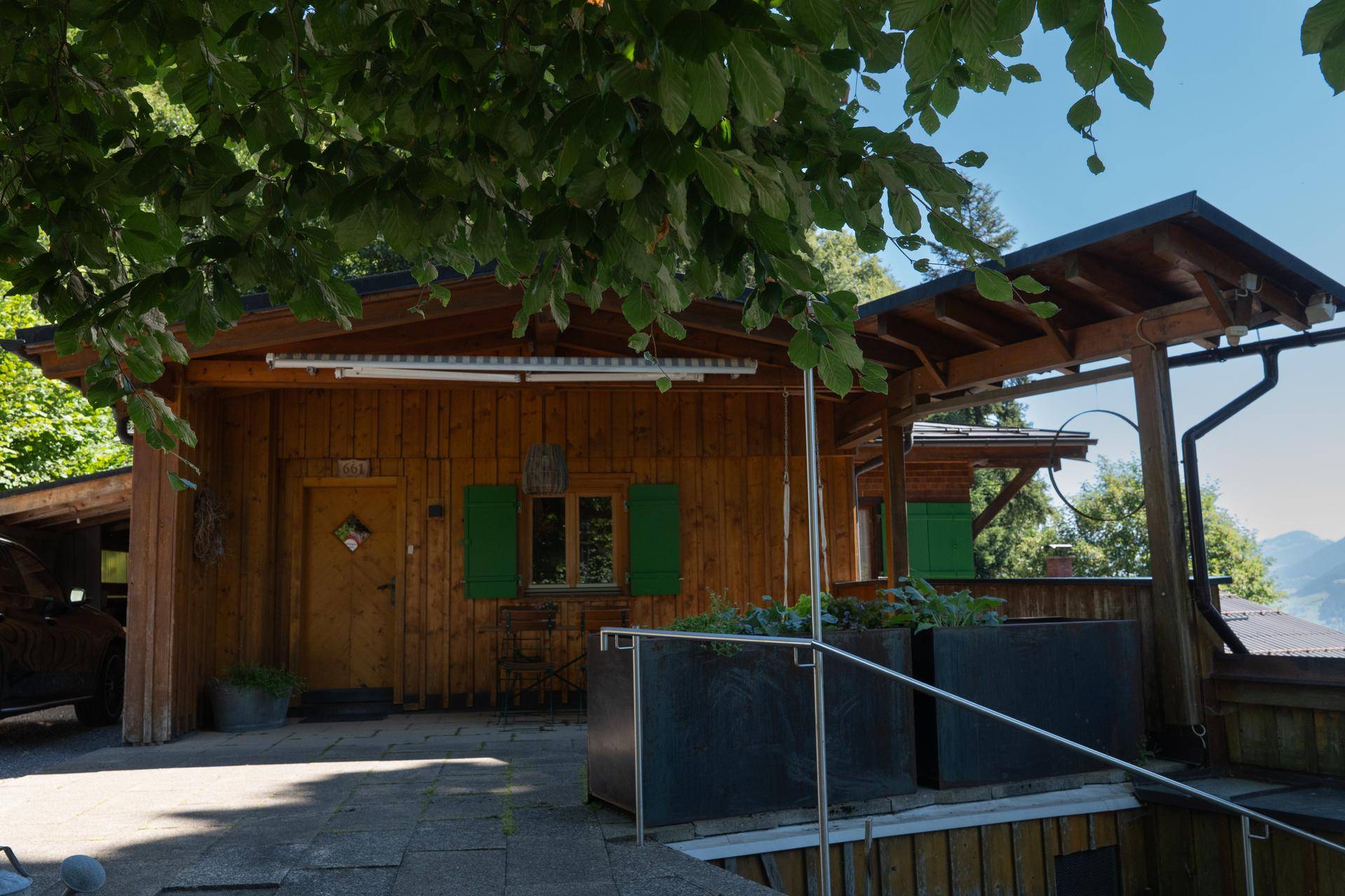Eingangsbereich des Holzchalets mit kleiner Terrasse und grünen Fensterläden unter schattigen Bäumen.