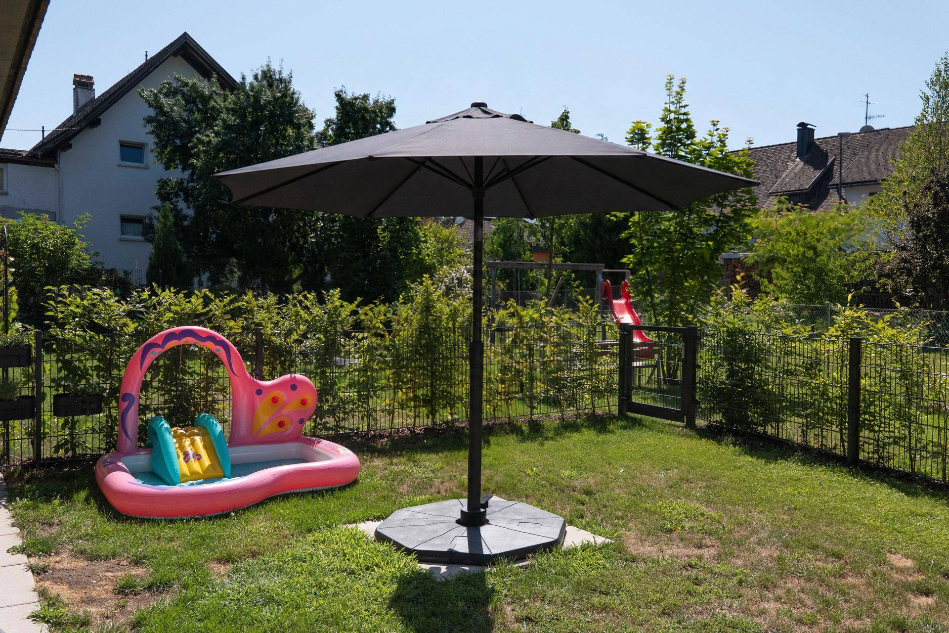 Sonniger Gartenbereich mit Planschbecken, Sonnenschirm und Spielgeräten im Hintergrund.