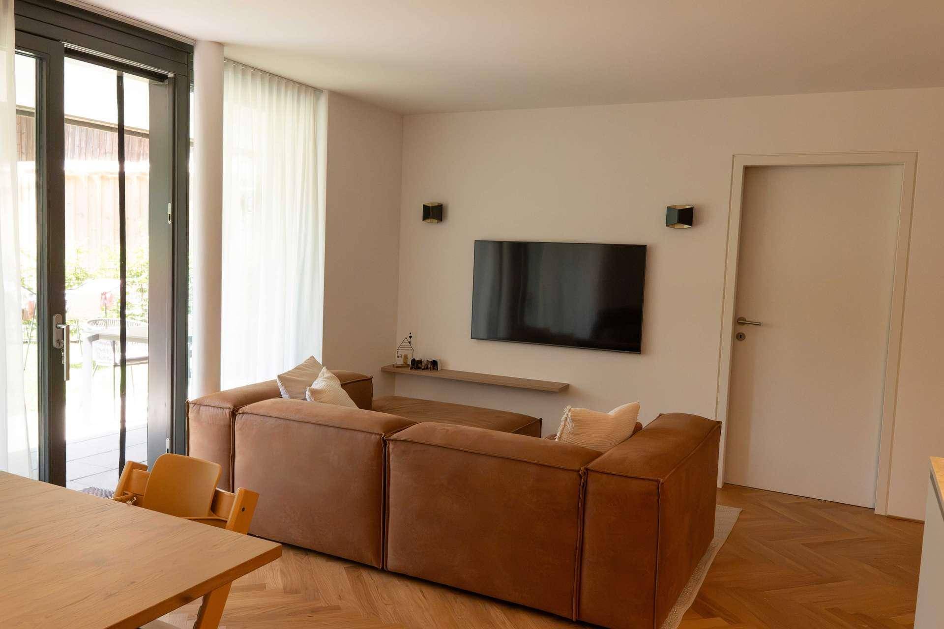 Gemütlicher Wohnbereich mit braunem Sofa, Fernseher und großen Fenstern zum Außenbereich.