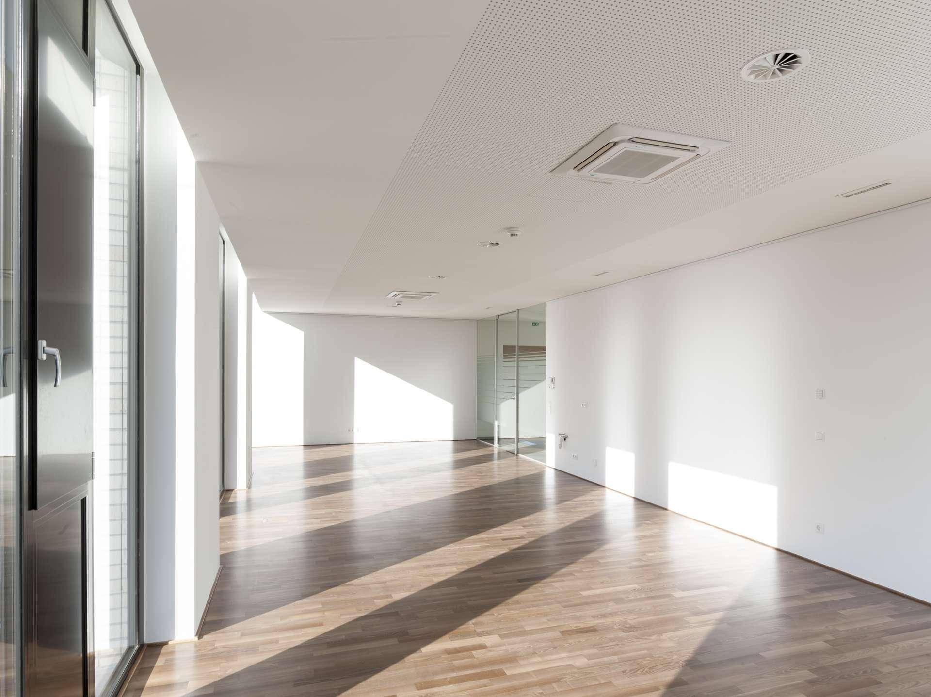 Heller Büroraum mit Holzboden, großen Fenstern und moderner Deckenlüftung, bereit zum Bezug.