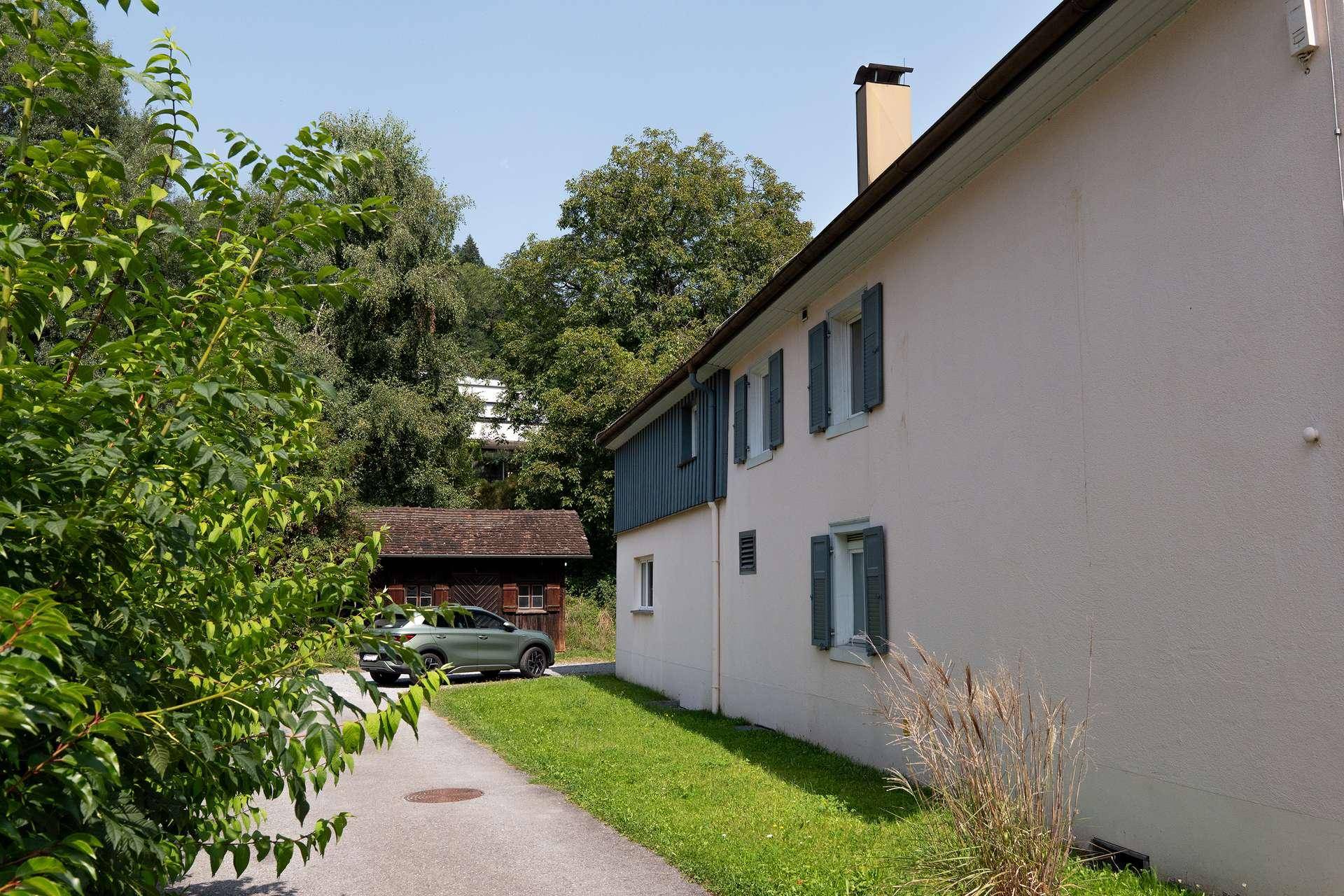 Seitenansicht eines Gebäudes mit blauen Fensterläden und einer Zufahrt, umgeben von üppigem Grün.