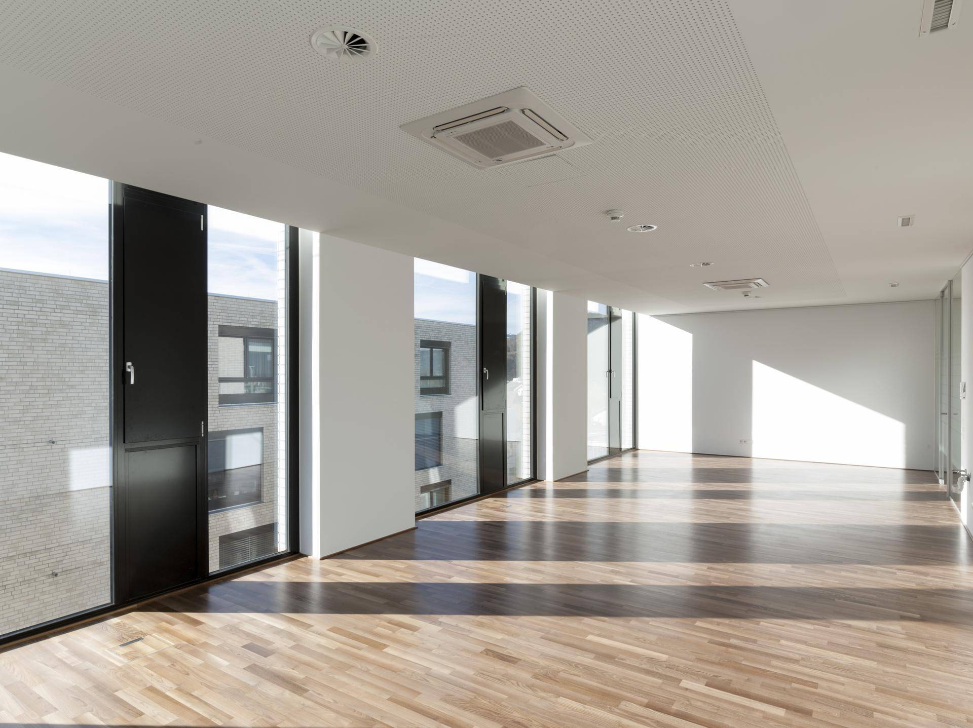 Heller und geräumiger Büroraum mit Holzboden und vielen Fenstern, die eine moderne und einladende Atmosphäre schaffen.