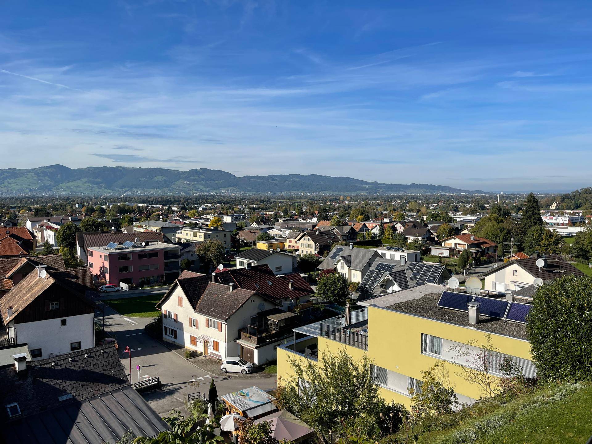 Weitläufiger Blick über eine Siedlung mit Häusern und Bergen im Hintergrund unter blauem Himmel.
