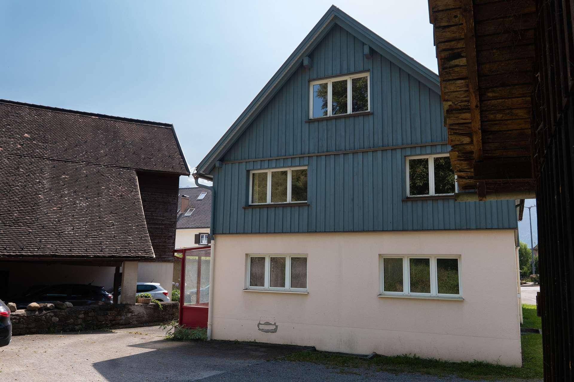 Seitenansicht eines Gebäudes mit blauer Holzverkleidung und hellen Wänden, angrenzend an einen Schuppen.
