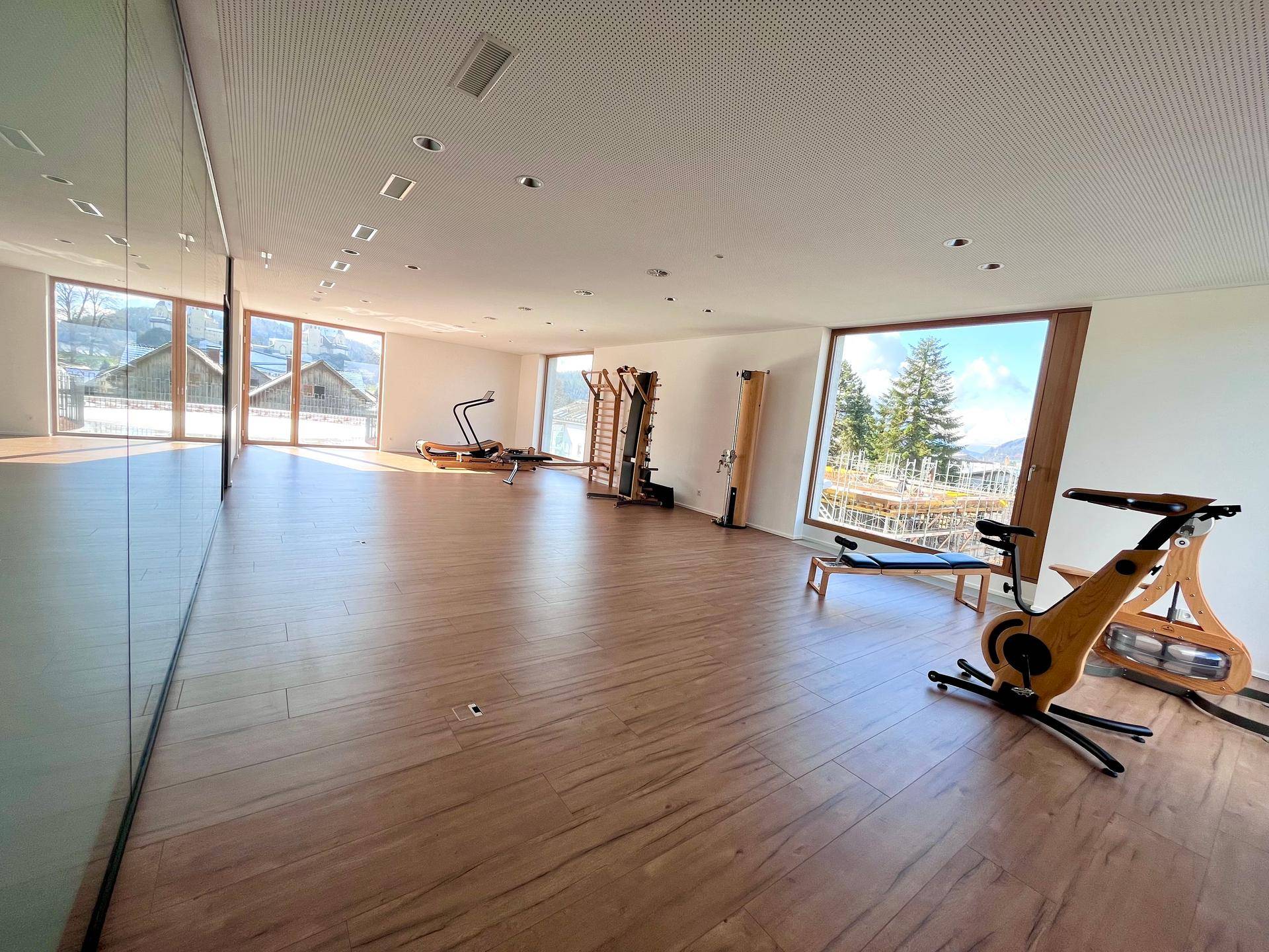 Großer Fitnessraum mit Trainingsgeräten, Holzboden und weitem Blick auf die umliegende Berglandschaft.