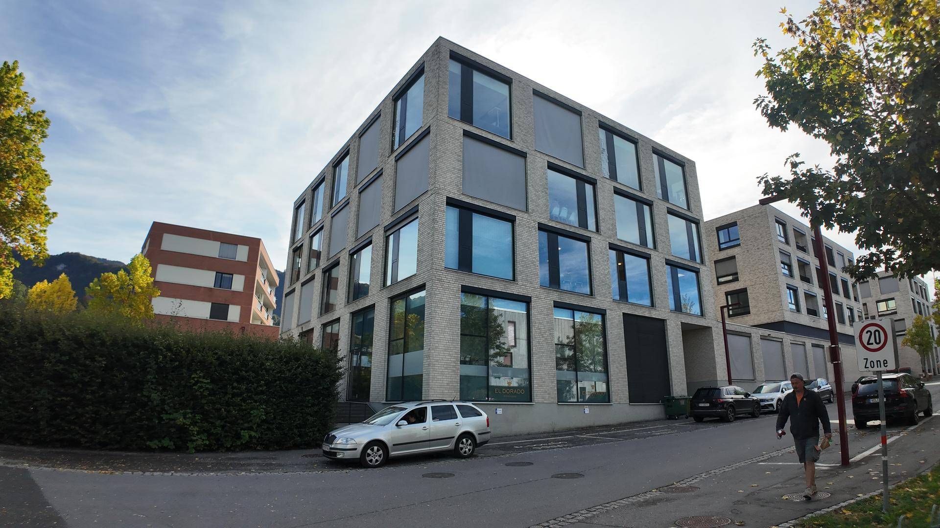Modernes Bürogebäude mit großen Fenstern und einer gepflegten Hecke an der Straße, umgeben von Parkplätzen.