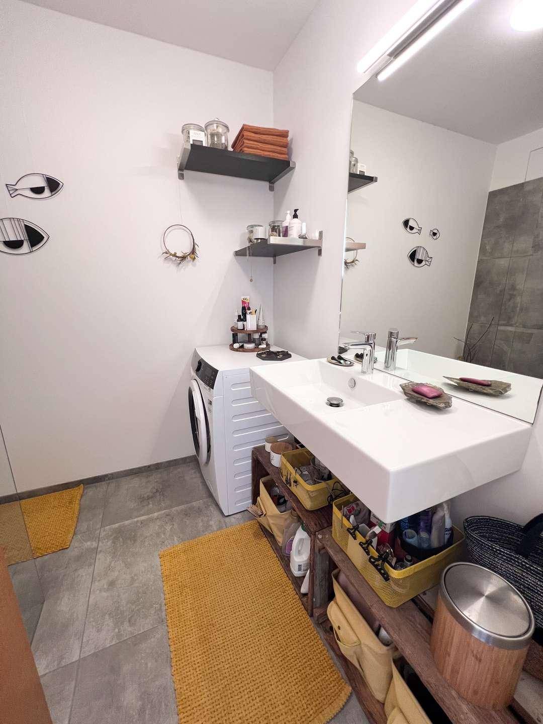Badezimmer mit Waschbecken, Spiegel, Waschmaschine und grauen Fliesen.