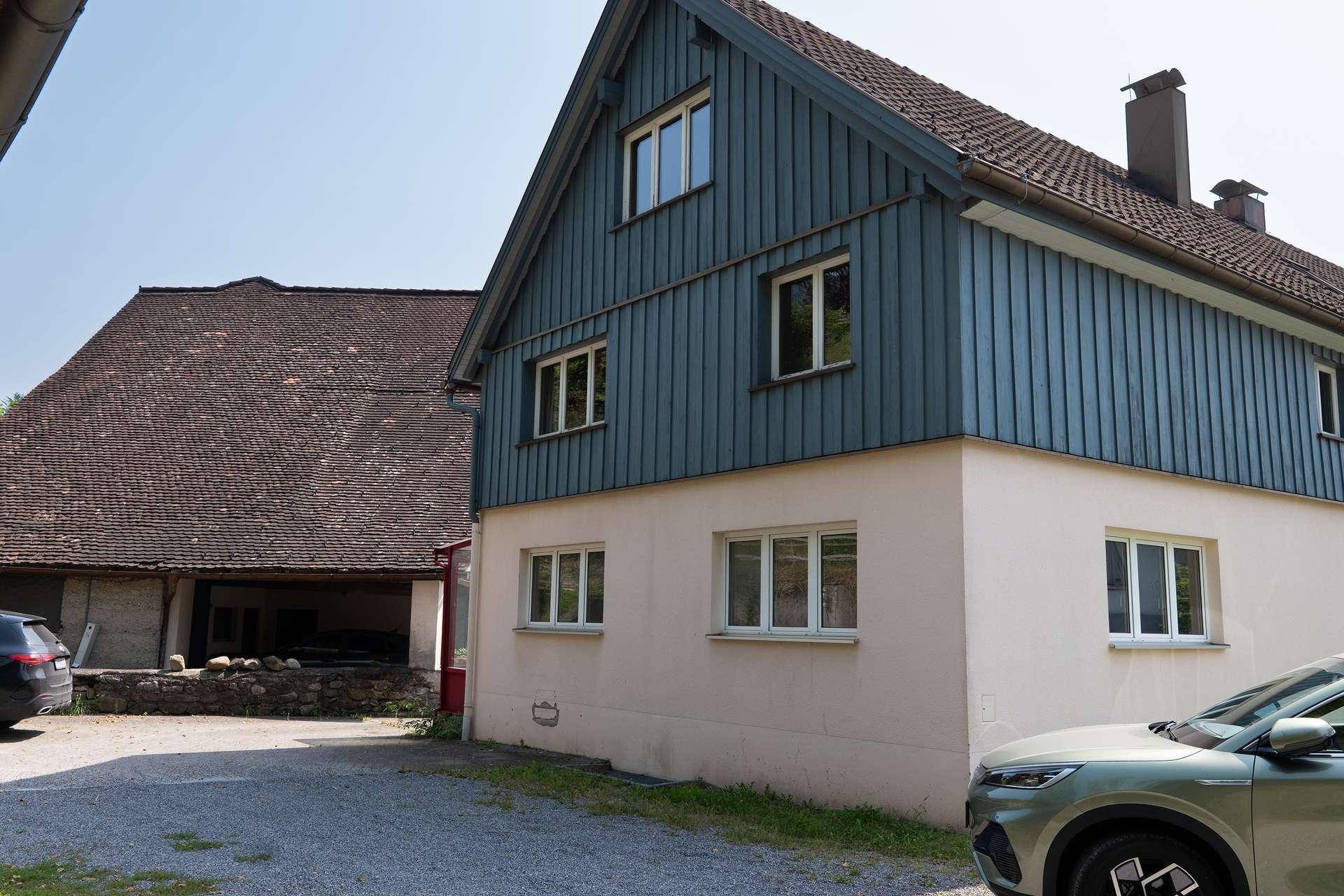 Seitenansicht eines Gebäudes mit blauer Holzverkleidung im oberen Bereich und hellen Fassaden im Erdgeschoss.