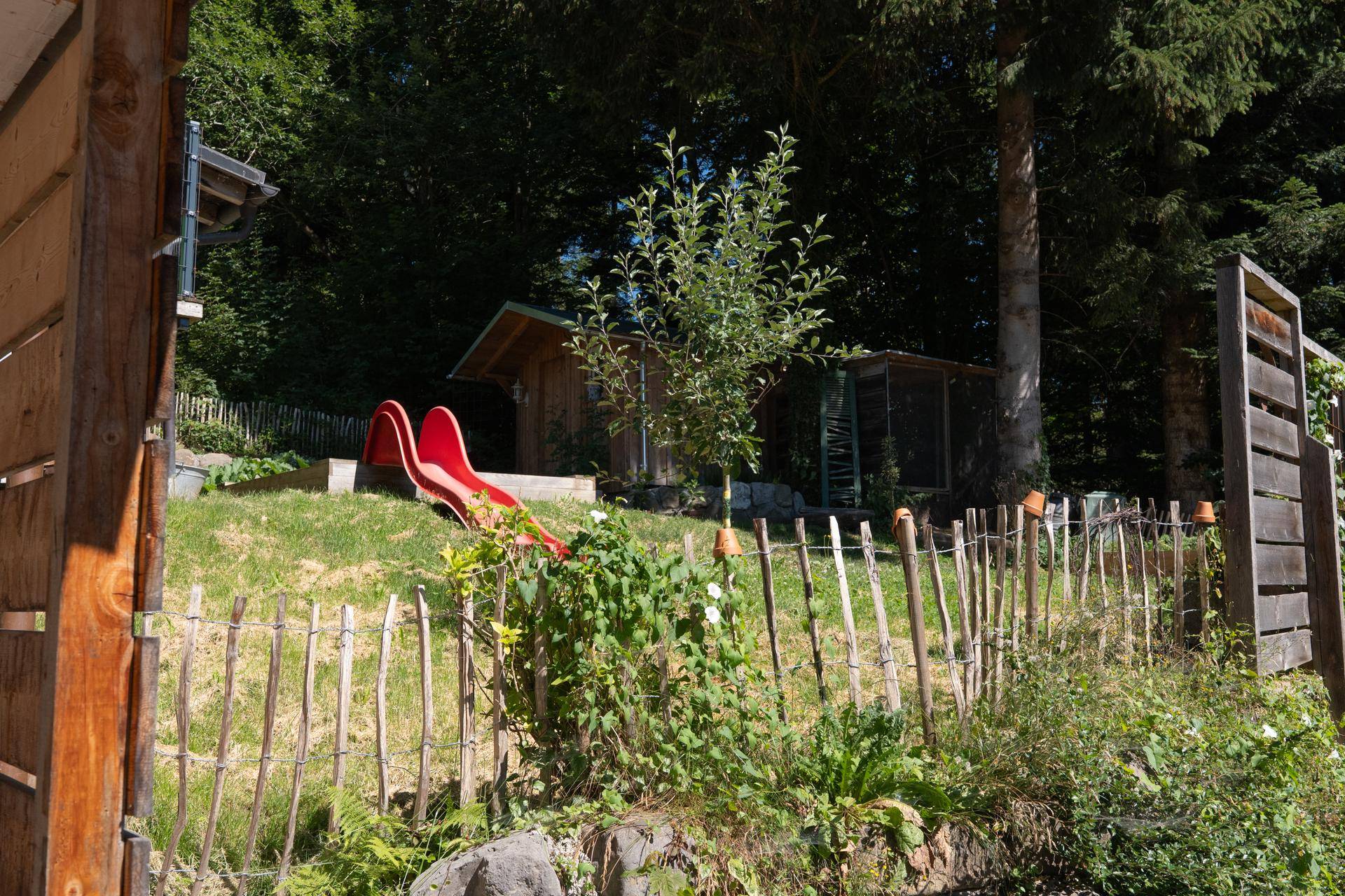 Spielbereich im Garten mit einer roten Rutsche, umgeben von Bäumen und einem rustikalen Zaun.