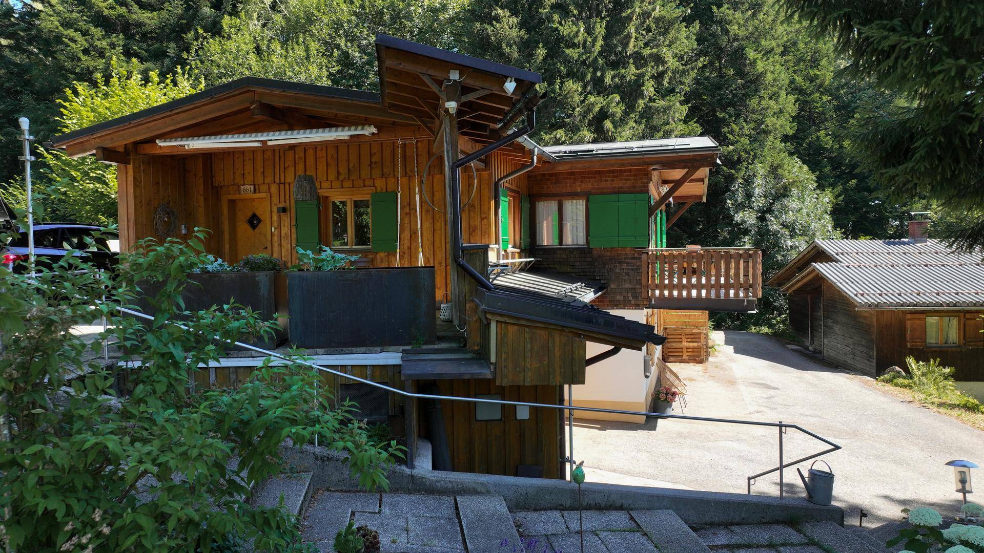 Seitenansicht des Holzhauses mit Außentreppe und mehreren Ebenen, umgeben von Grün.