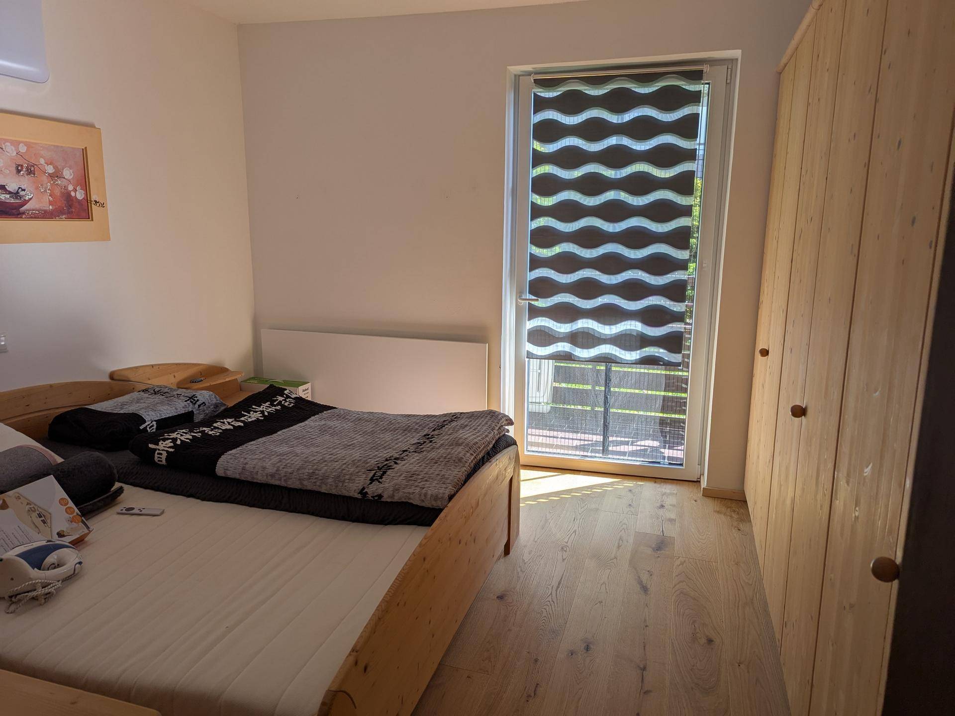 Schlafzimmer mit Holzbett, Holzschrank und Zugang zu einem Balkon mit Rollo.