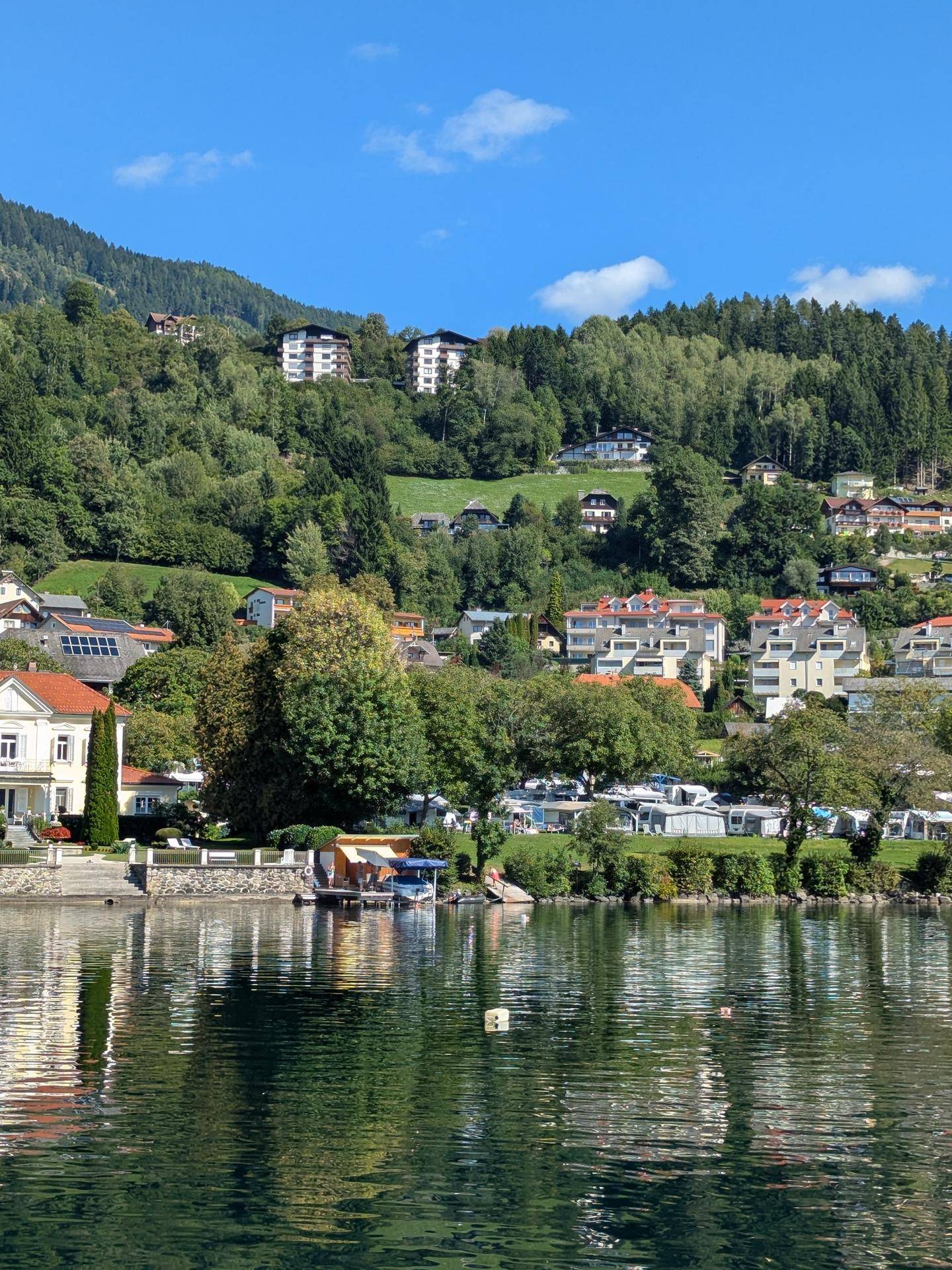 Malerische Ansicht des Sees mit Häusern und grünen Hügeln im Hintergrund unter blauem Himmel.
