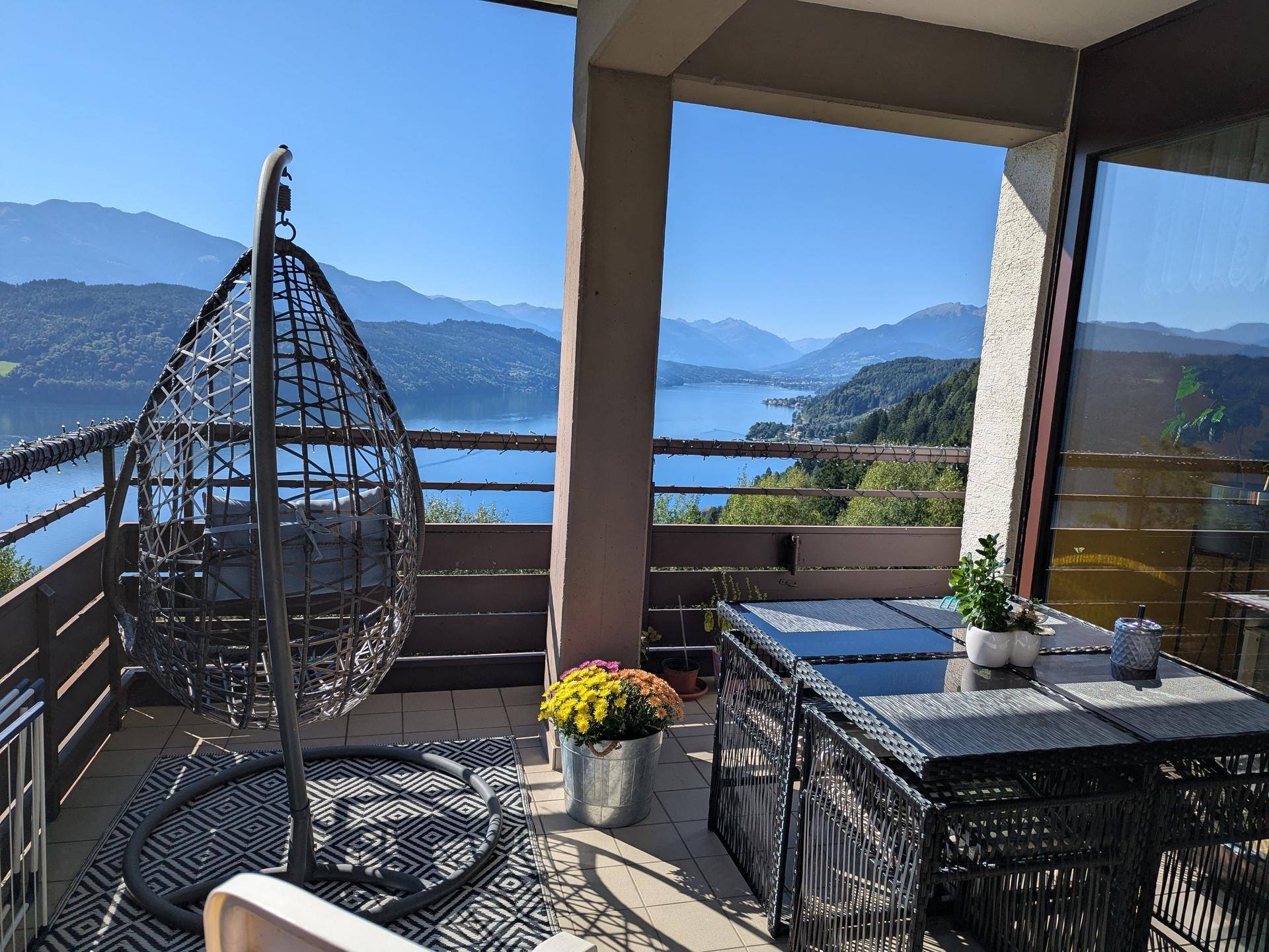 Möblierter Balkon mit Hängesessel und Esstisch, bietet einen atemberaubenden Blick auf den See und die Berge.