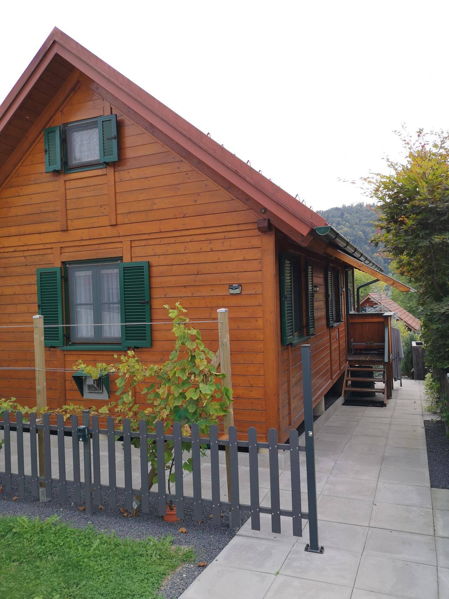 Seitenansicht eines Holzhauses mit grünen Fensterläden und einem gepflasterten Weg.
