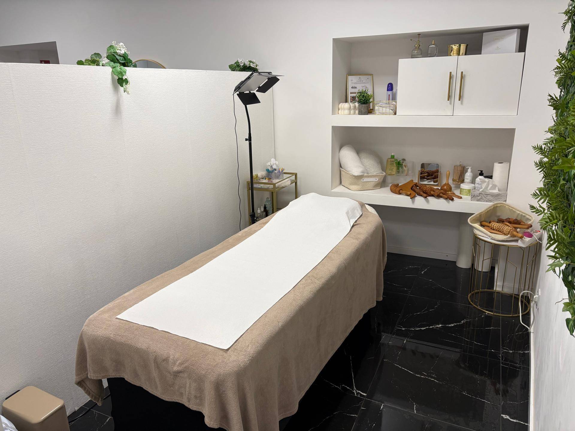 Behandlungsraum mit Liege, weißer Bettwäsche und integrierten Wandregalen für Produkte.