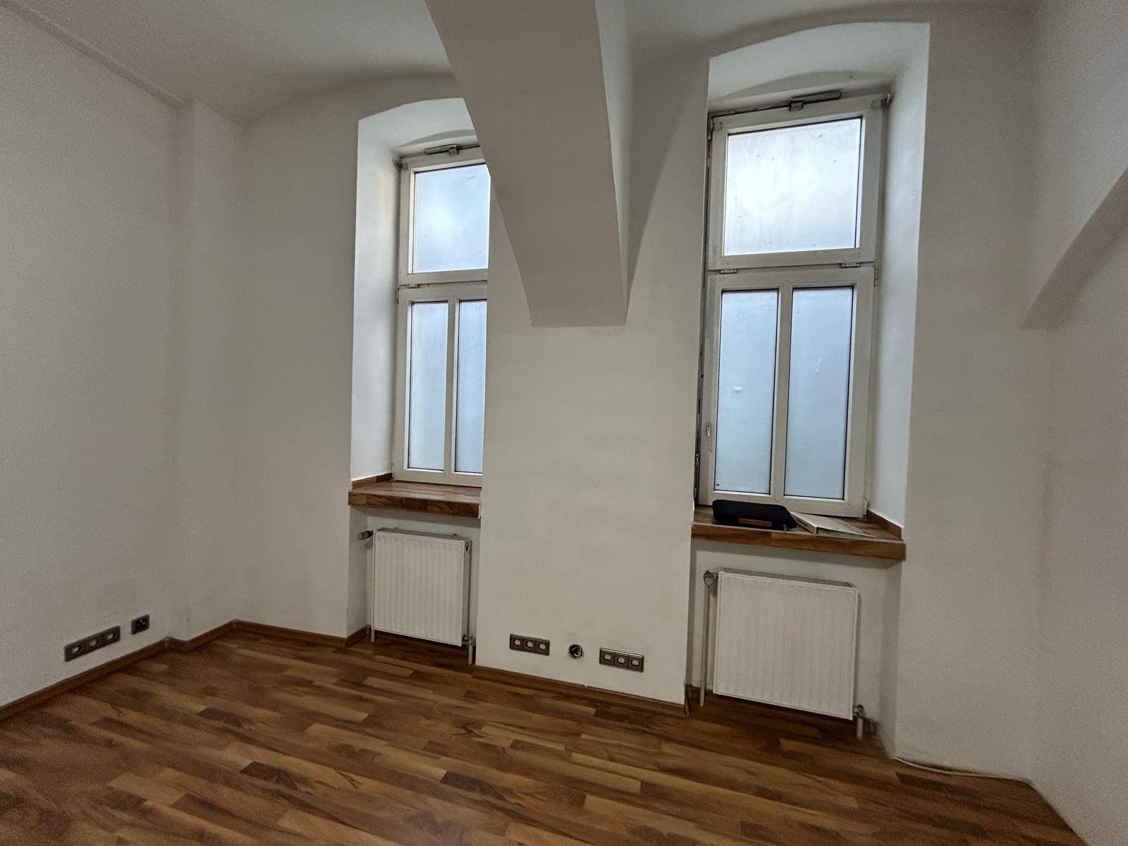 Heller Raum mit zwei hohen Fenstern, Laminatboden und Heizkörpern unter den Fensterbänken.