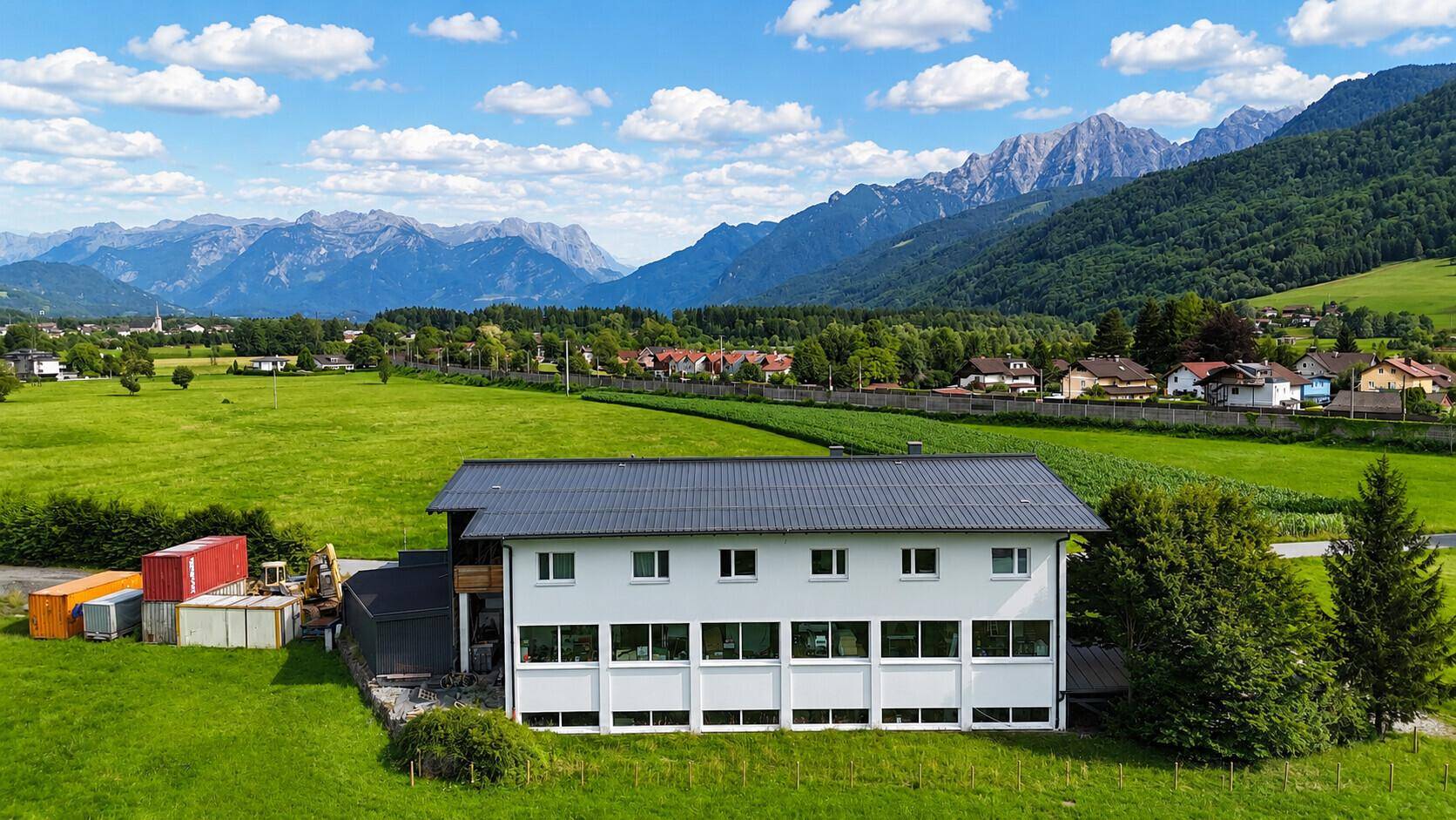 Rückansicht eines modernen Hauses mit großen Fenstern, umgeben von weiten grünen Feldern und Bergpanorama.