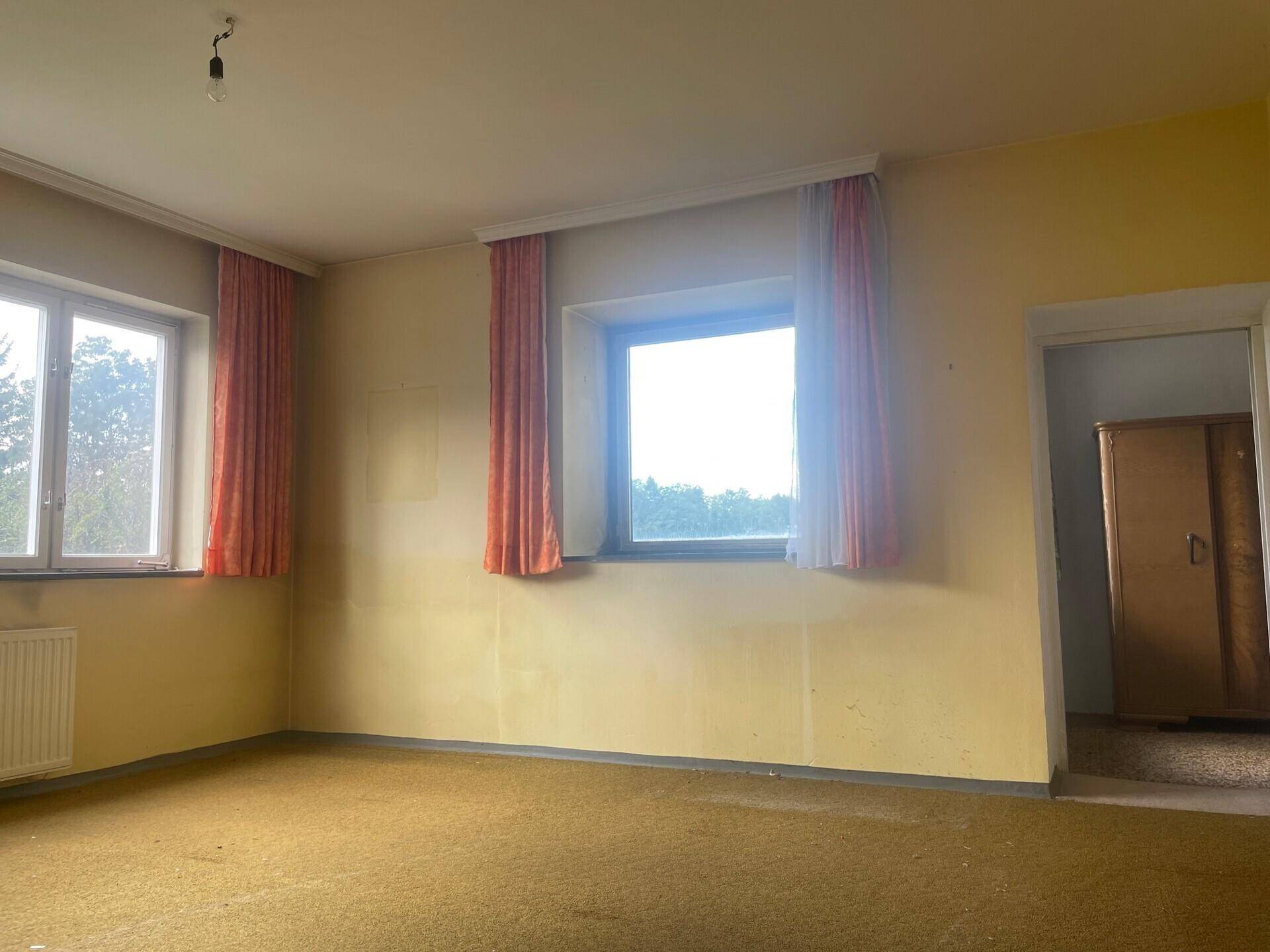 Leeres Zimmer mit gelben Wänden, Teppichboden und zwei Fenstern mit Vorhängen.
