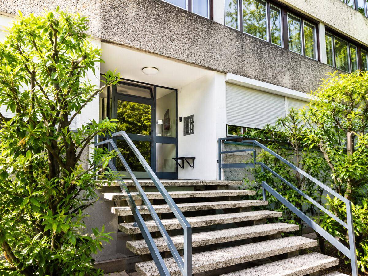 Eingangsbereich eines Wohnhauses mit Steintreppe und Geländer, umgeben von Grün.