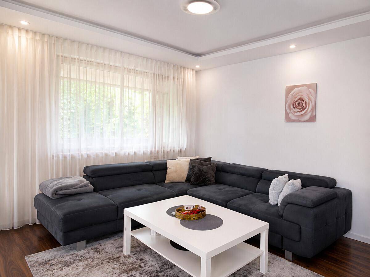 Gemütliches Wohnzimmer mit großem Ecksofa, hellem Teppich und Vorhängen am Fenster.
