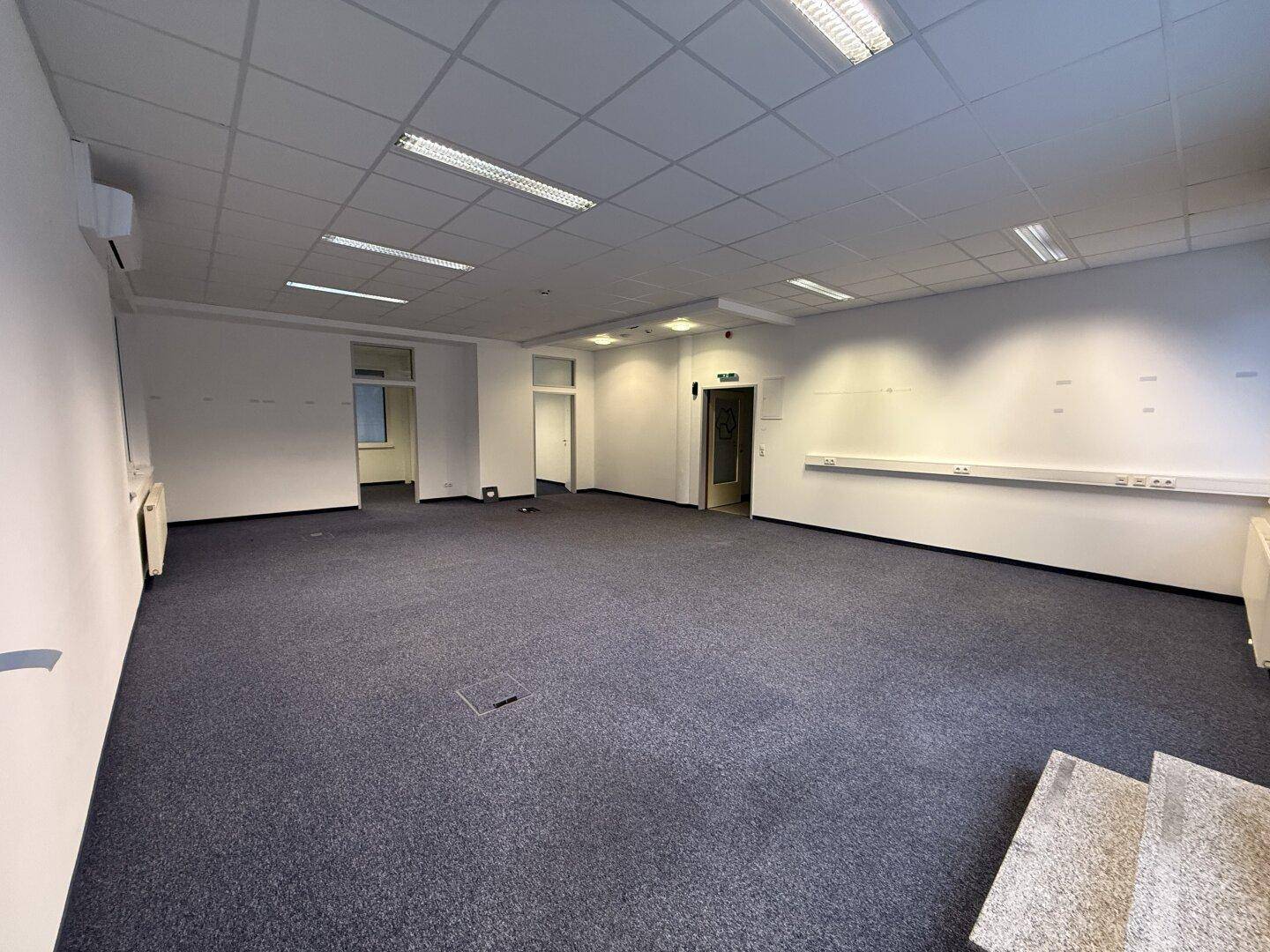 Weitläufiges, leeres Büro mit Teppichboden, Systemdecke und Zugang zu weiteren Räumen im Hintergrund.