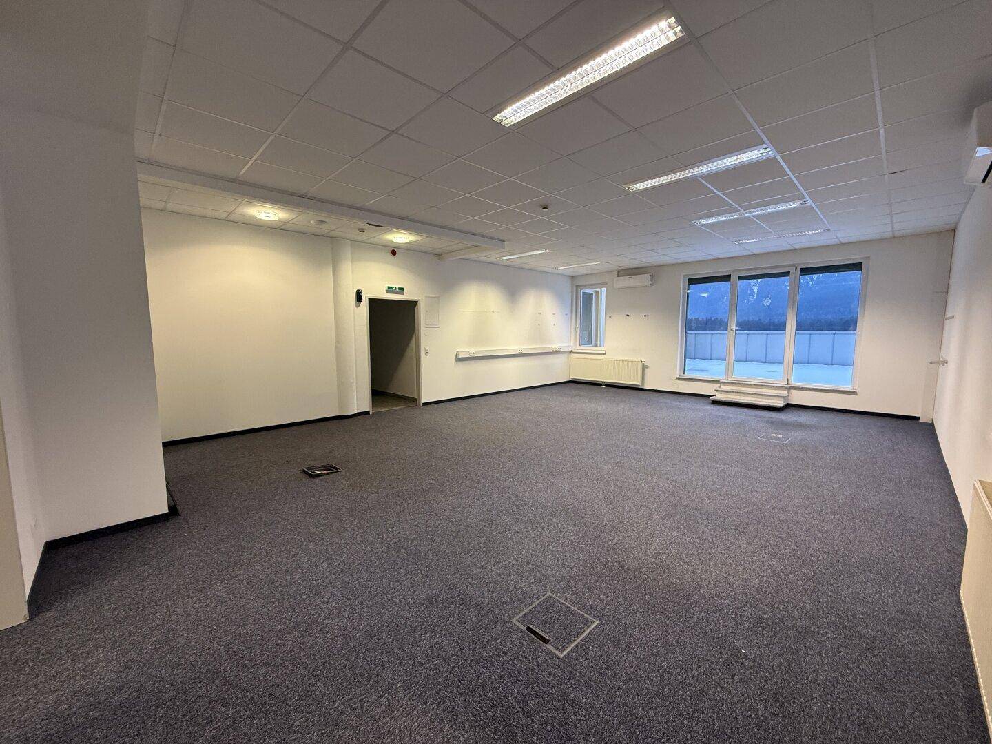 Helles, leeres Büro mit Teppichboden, abgehängter Decke und Blick auf die Berge durch die Fenster.