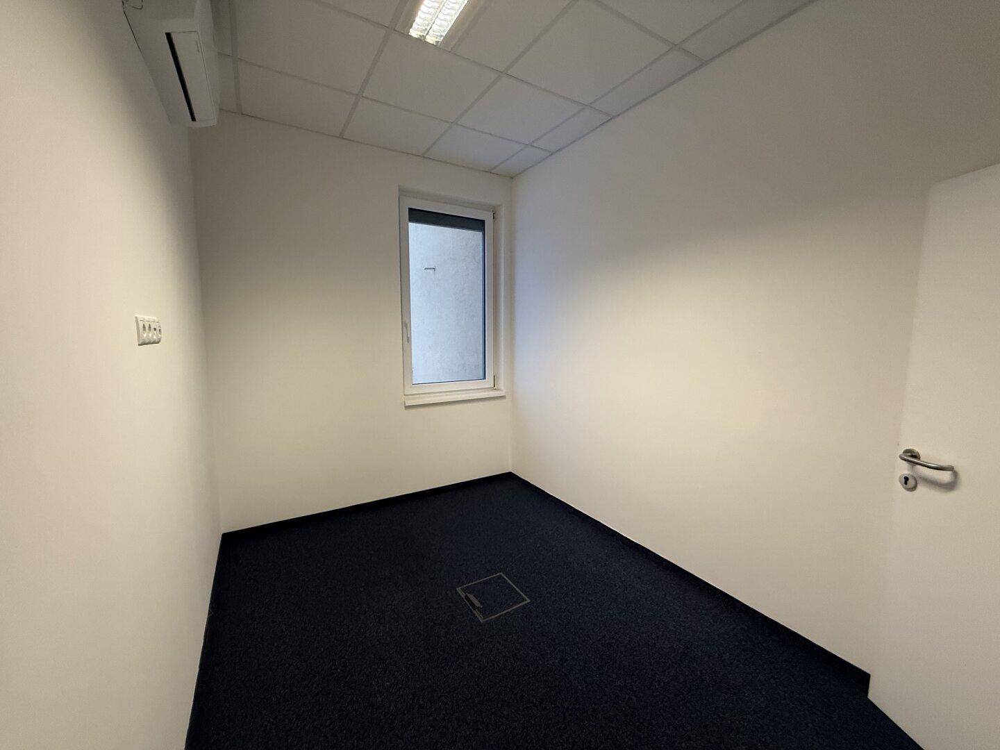 Kompaktes Büro mit Teppichboden, einem Fenster und Klimaanlage, ideal für konzentriertes Arbeiten.