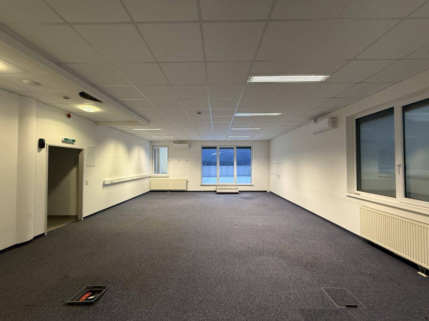 Geräumiges, leeres Büro mit Teppichboden, abgehängter Decke und großen Fenstern mit Blick nach draußen.