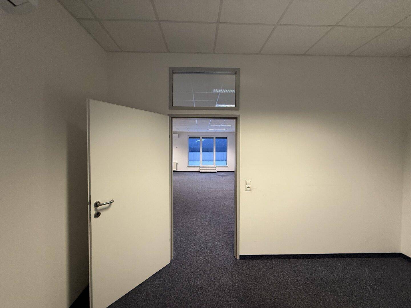 Büro mit offener Tür, die den Blick in einen größeren, hellen Büroraum freigibt.