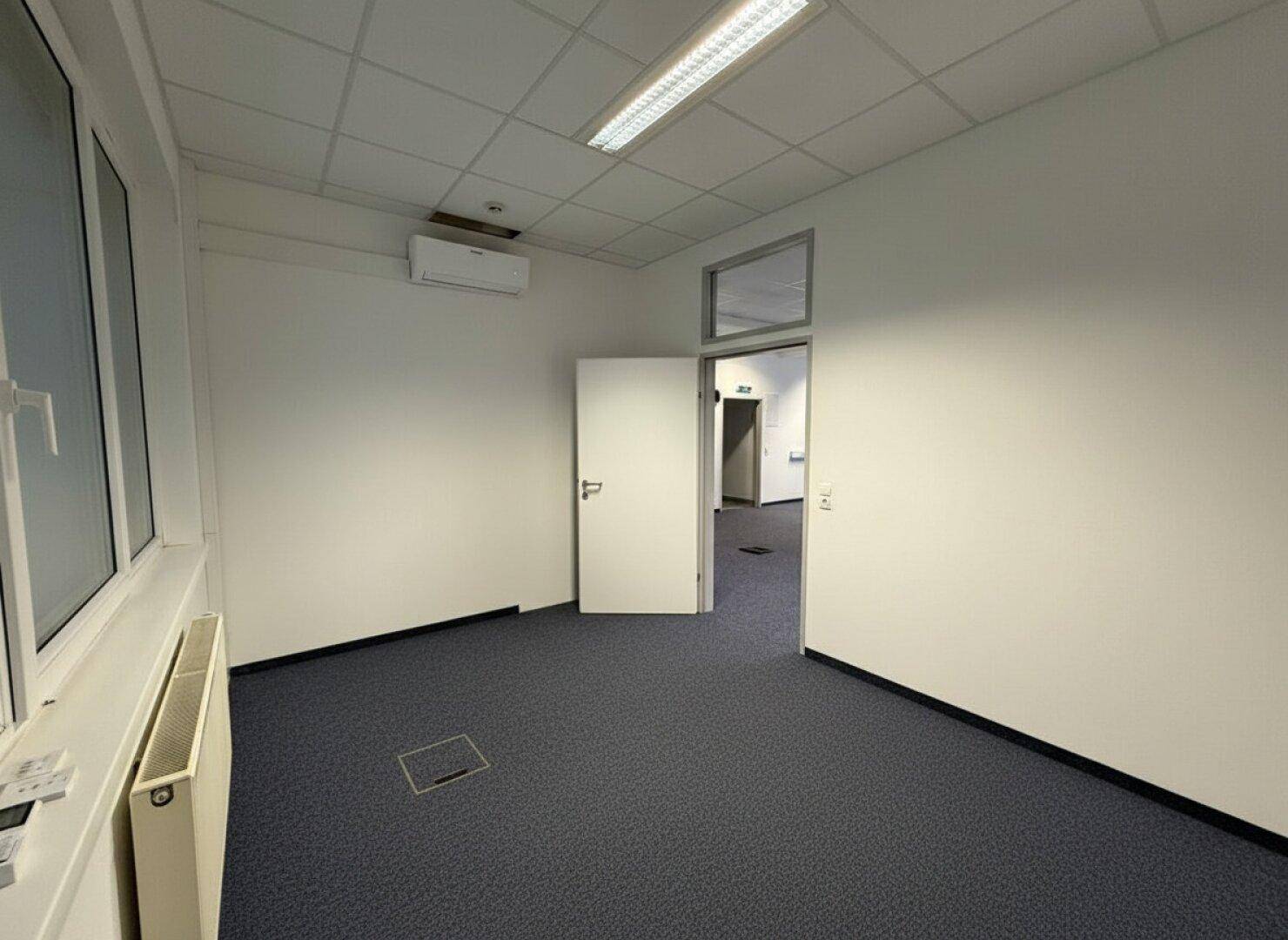 Einzelbüro mit Teppichboden, Fenster und offener Tür zu einem größeren Bürobereich.