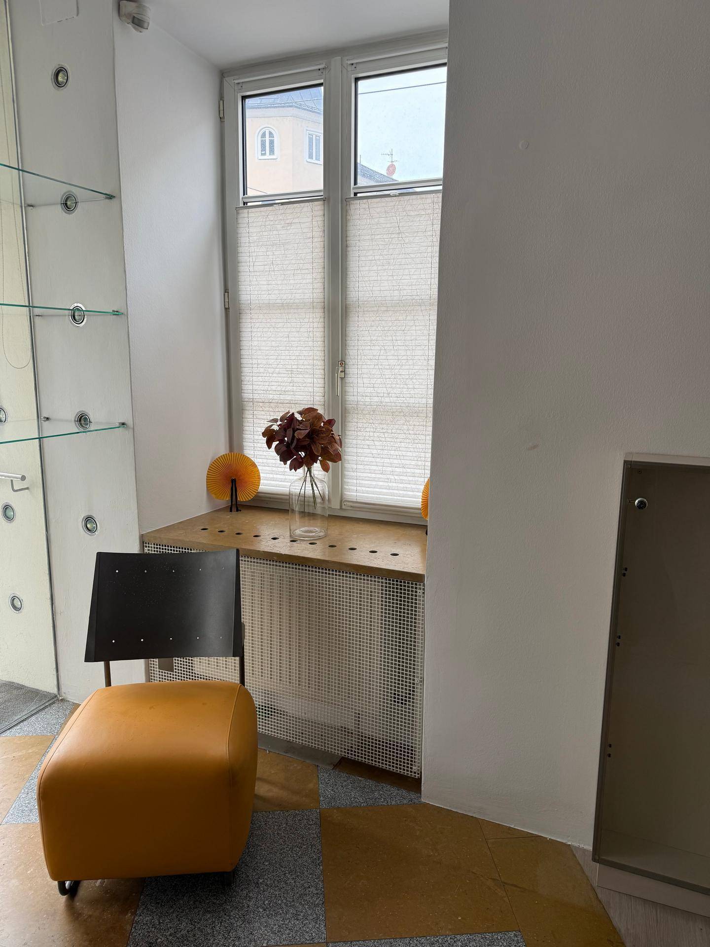 Fensterbereich mit einem Heizkörpergitter, einer Ablagefläche und einem gelben Hocker in einem Wartebereich.