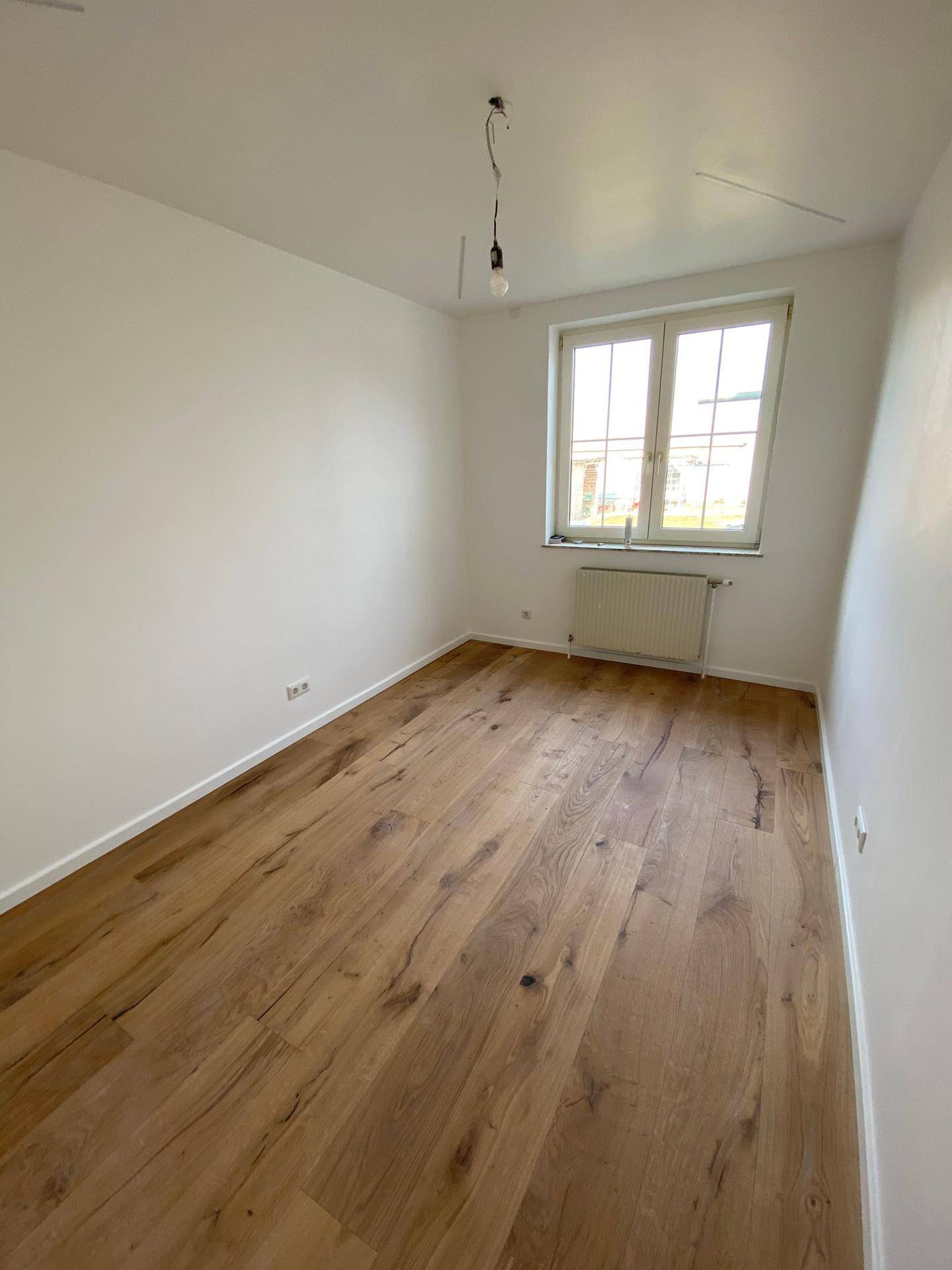 Helles Schlafzimmer mit Holzdielenboden, Heizkörper unter dem Fenster und weißen Wänden.