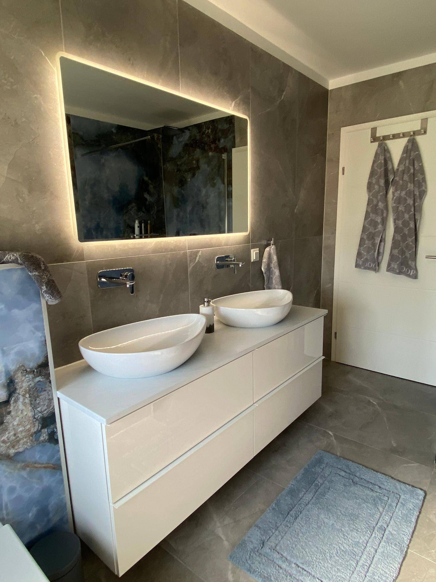 Modernes Badezimmer mit Doppelwaschbecken, beleuchtetem Spiegel und grauen Marmorfliesen.