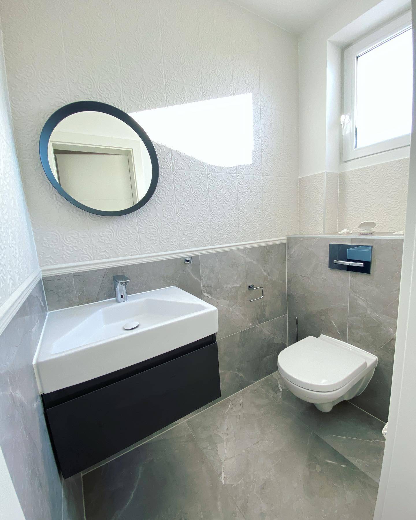 Modernes Gäste-WC mit grauen Fliesen, wandhängendem WC und kleinem Waschbecken mit Spiegel.